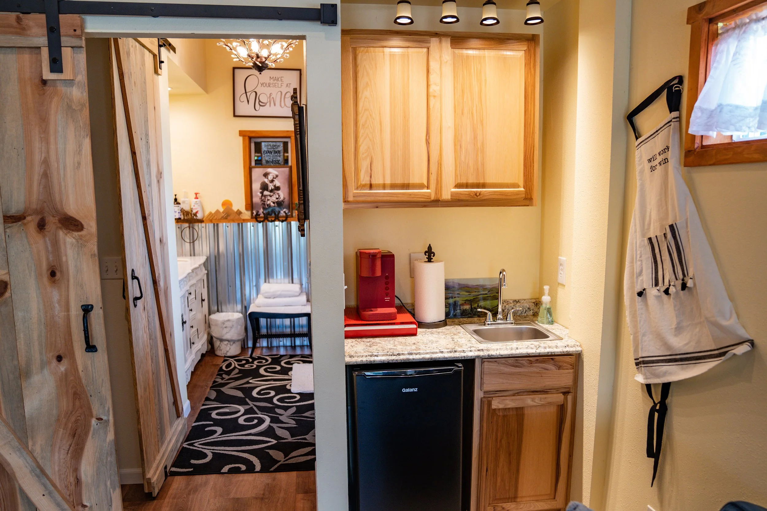 A small kitchen area with wooden cabinets and a granite countertop, featuring a red coffee maker, a paper towel dispenser, and a sink. To the right, a window with a white curtain, and on the wall, a hanging apron. Through an open doorway, a bathroom 