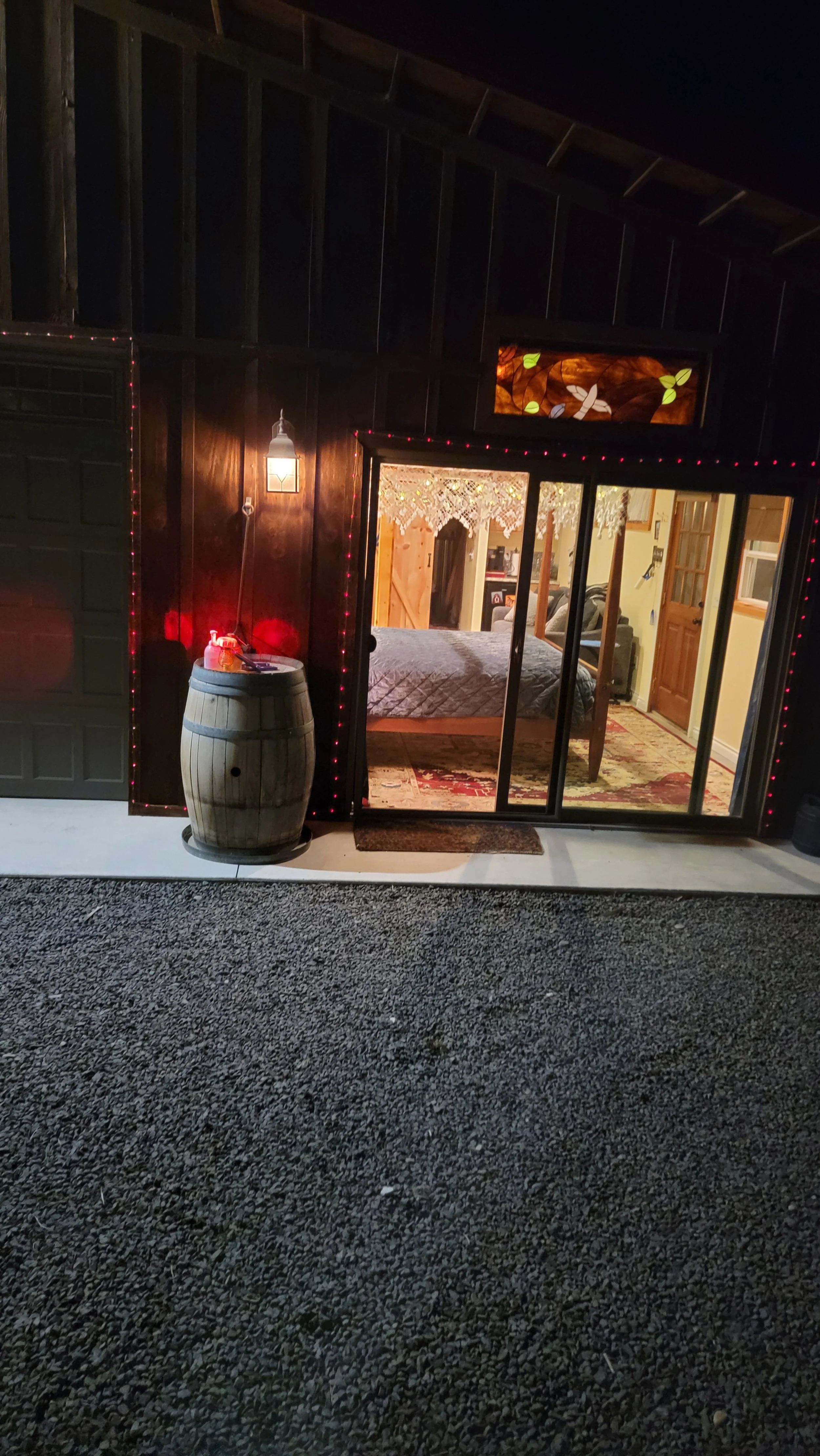 View of a cozy room inside a barn or workshop, seen through a sliding glass door, with warm lighting and rustic decor including a barrel, a bed, and string lights.