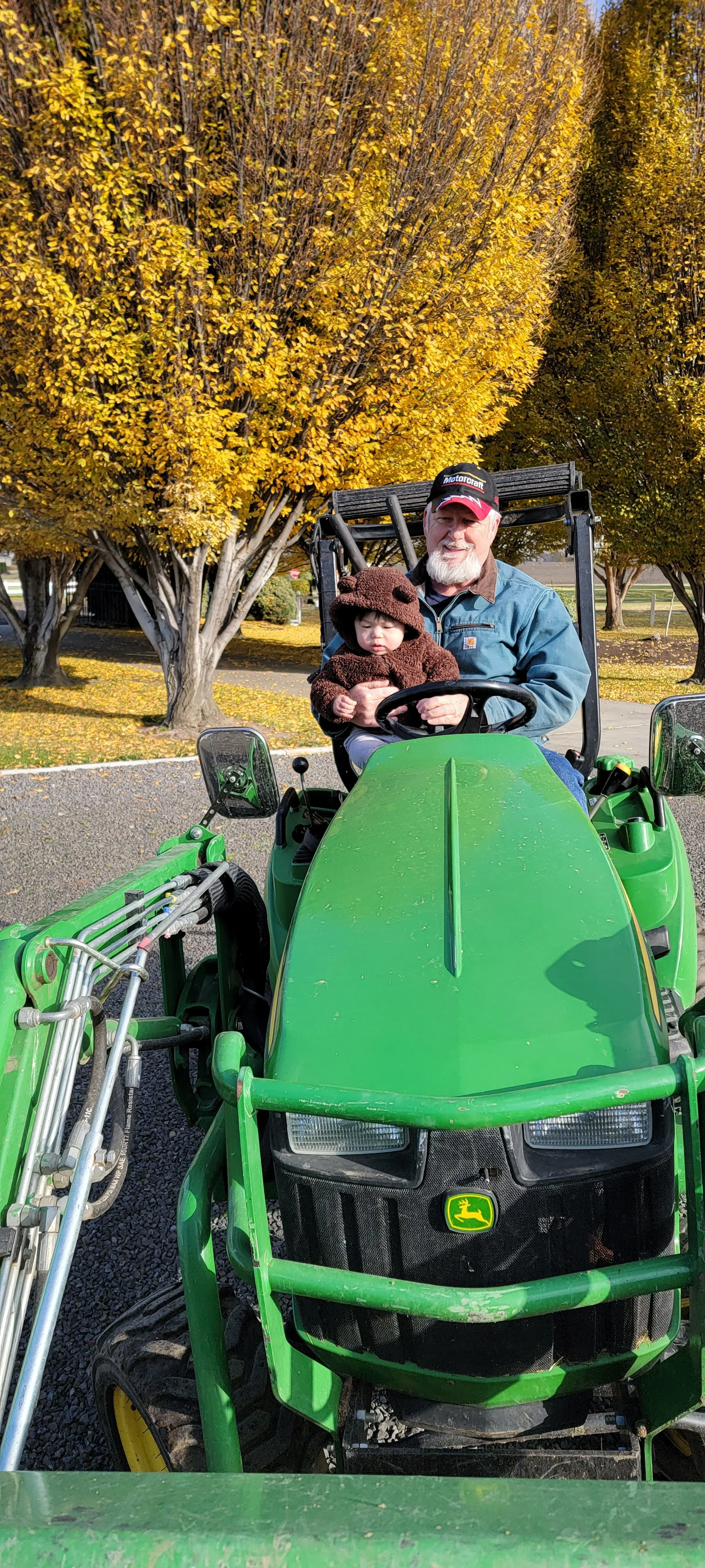 A man and a young child sitting on a green John Deere tractor in a park with fall foliage. The man has a beard, is wearing a cap and jacket, and is smiling. The child is dressed in a bear costume with bear ears and a face on the hood.