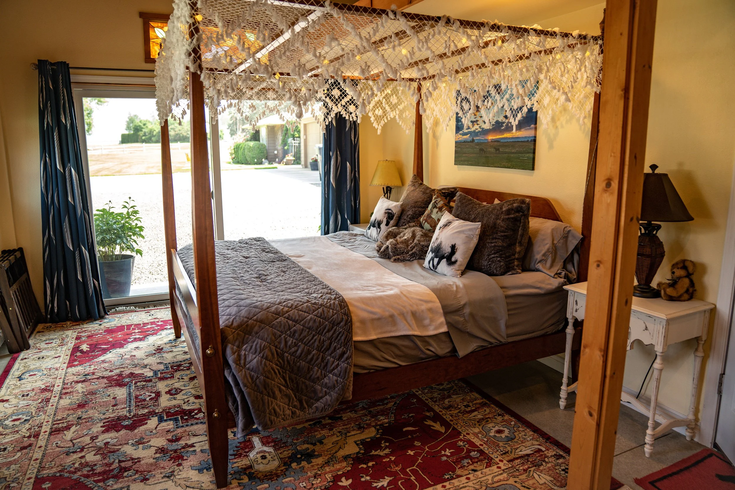 A bedroom with a wooden four-poster bed, a variety of pillows, and a blanket. There is a hanging canopy made of white fabric above the bed. The room has sliding glass doors with blue curtains, leading outside to a driveway and yard. The room features