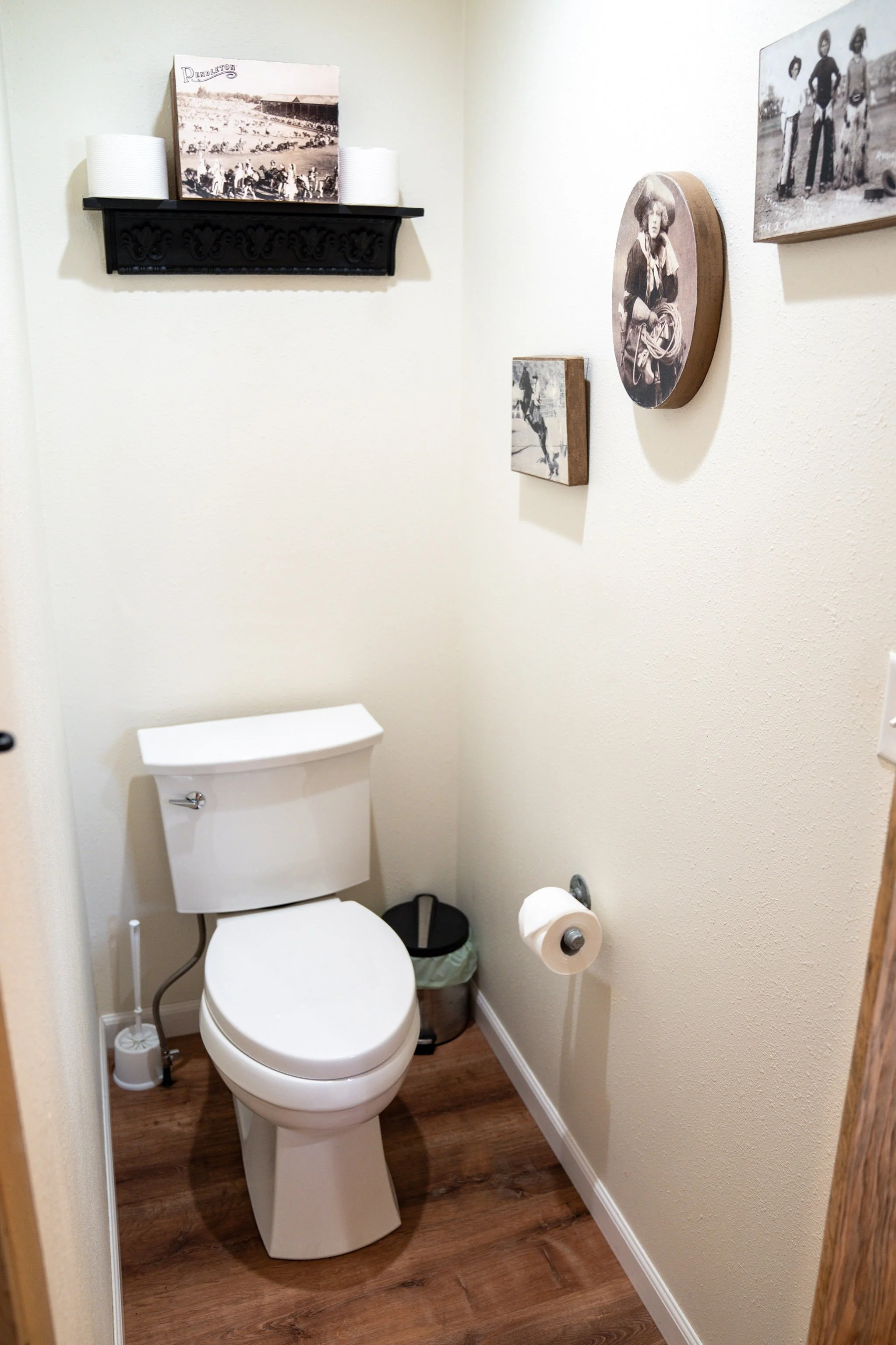 A small bathroom with a white toilet, a toilet brush, and custom framed black-and-white photos and artwork on the walls.