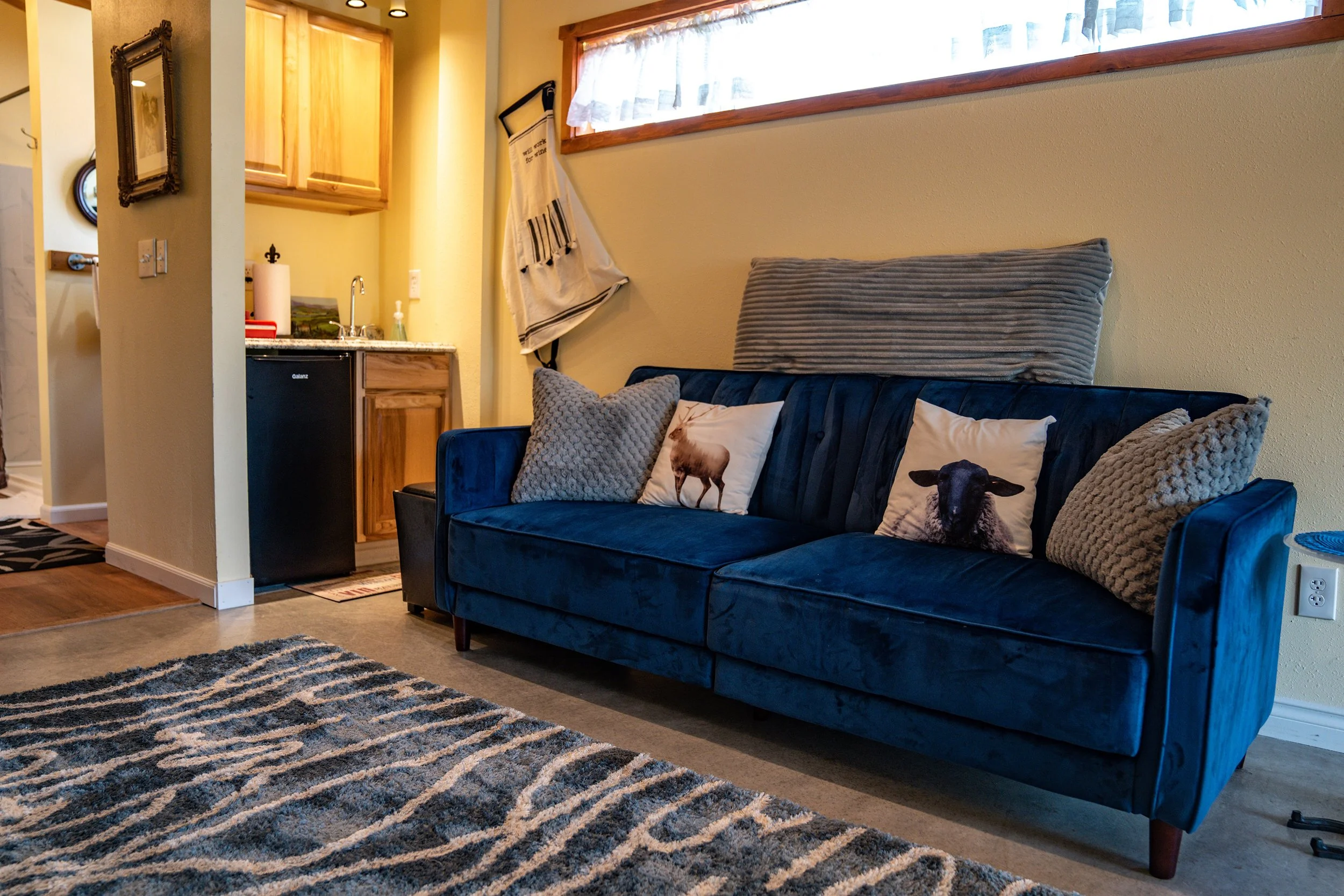 A cozy living room with a blue velvet sofa decorated with pillows featuring farm animal prints and textured pillows, adjacent to a small kitchenette with wooden cabinets, a mini-fridge, and a window with lace curtains.