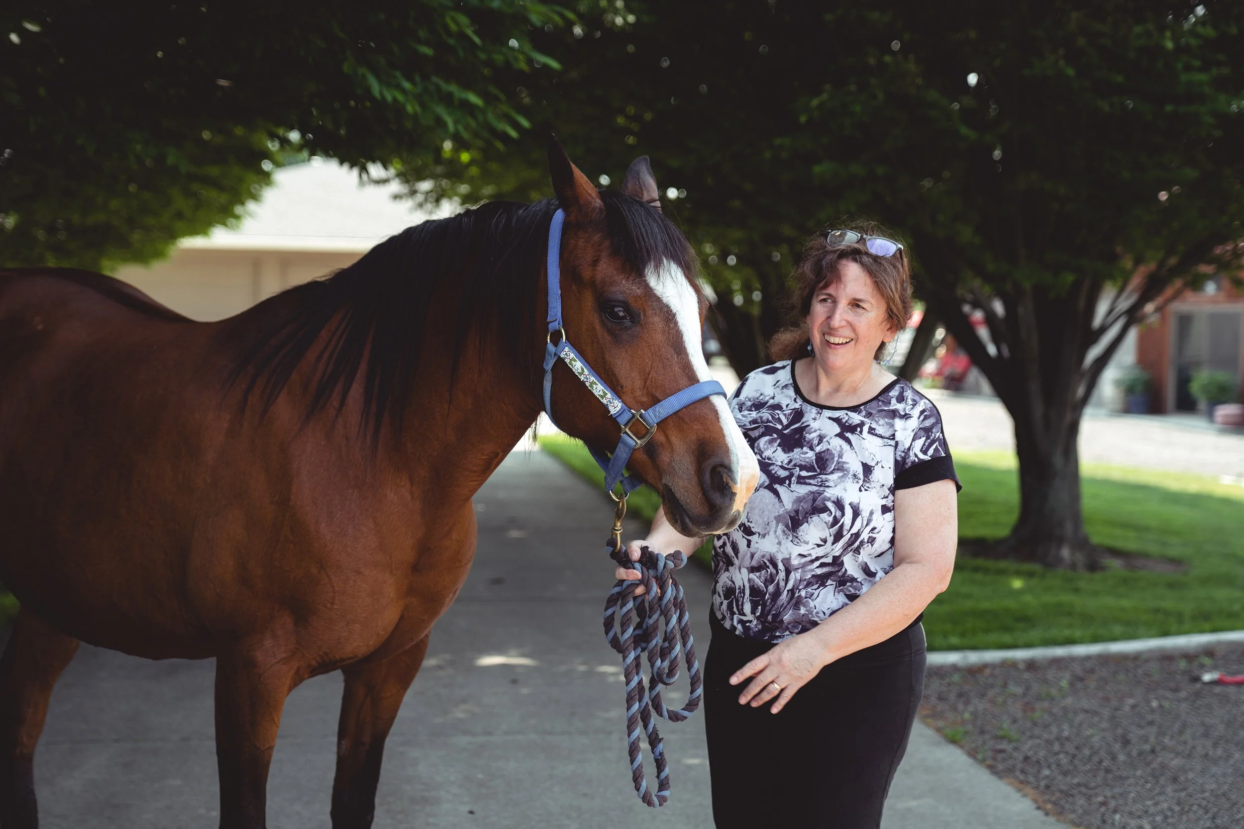 A woman smiling and holding a blue rope lead attached to a brown horse with a white blaze on its face, standing under the shade of trees in a park or driveway.