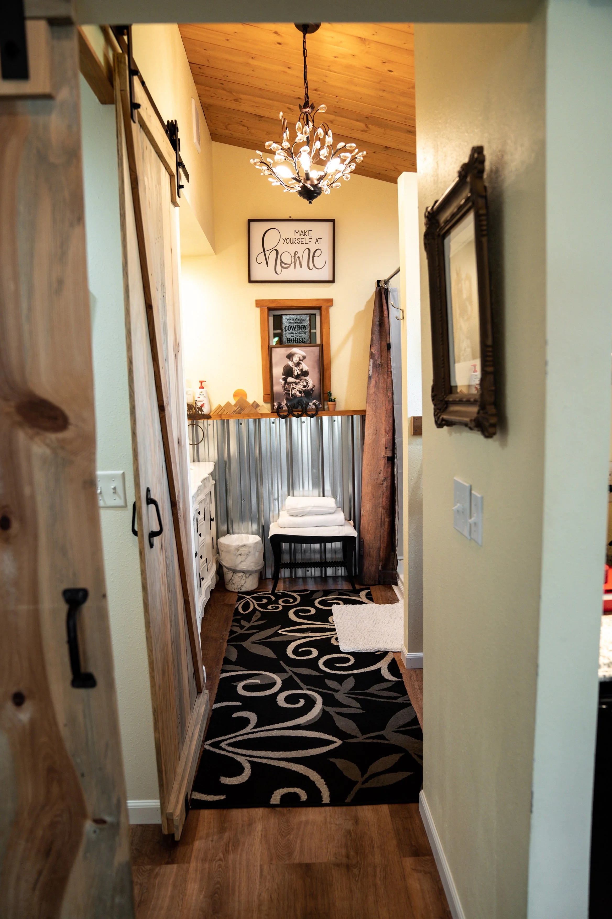 A cozy entryway with a wooden ceiling, chandelier, framed wall art that reads 'Make Yourself at Home,' a small bench with folded towels, a decorative rug with swirling patterns, and various rustic decor items.