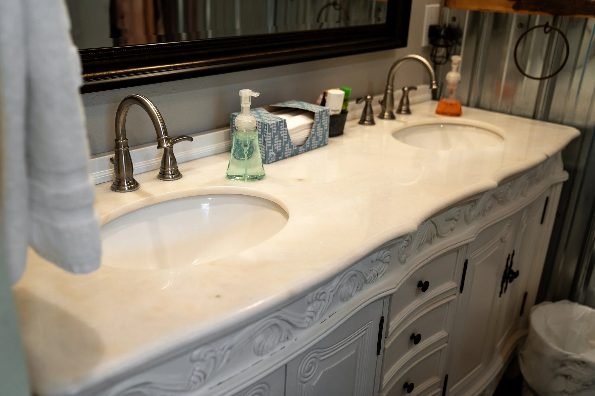 A bathroom vanity with two sinks, a marble countertop, and decorative cabinetry. There are soap dispensers, a tissue box, and other toiletries on the counter.