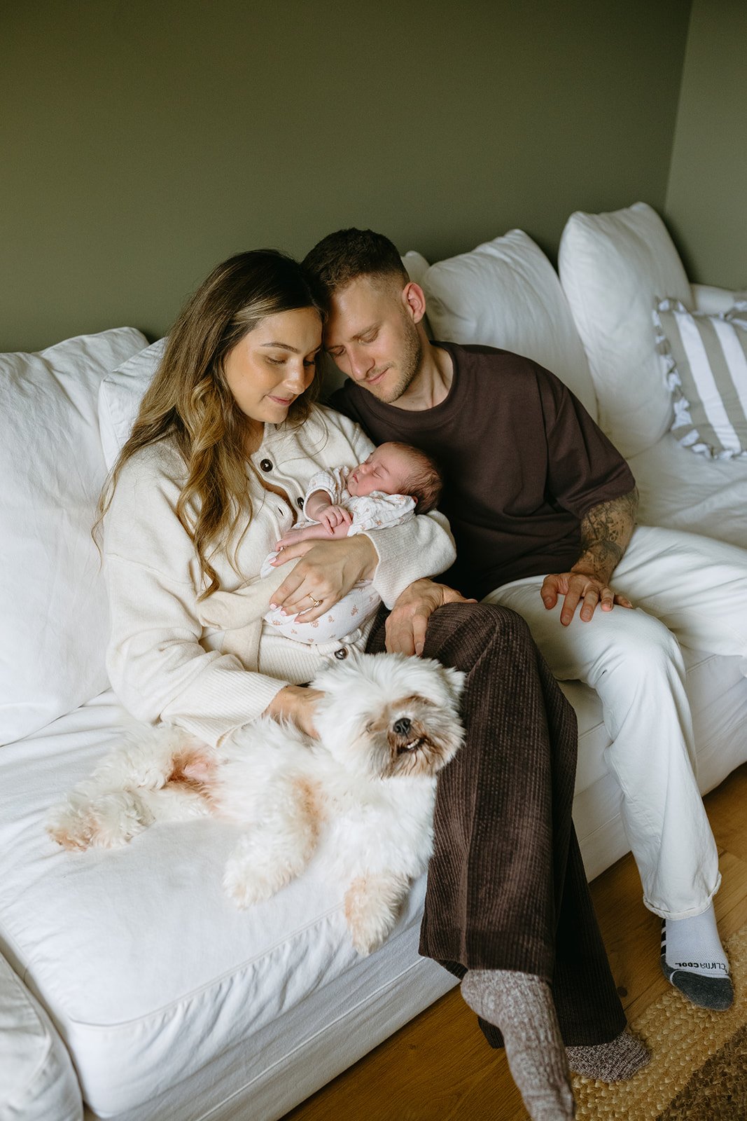 A family of three sitting on a white sofa with their dog, a woman holding a newborn, a man leaning in close, in a cozy living room.