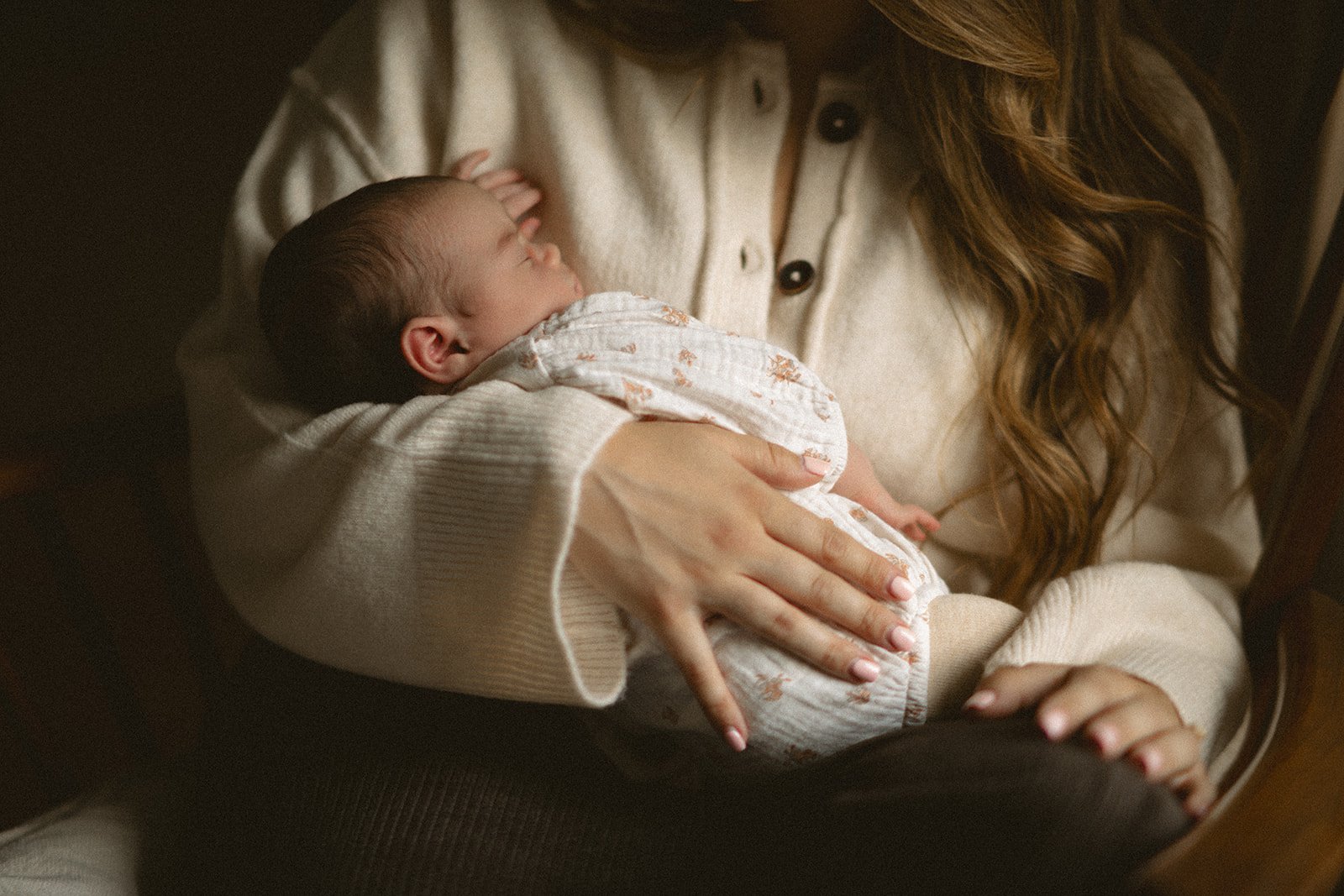 A woman holding a sleeping baby on her lap, with her hand gently resting on the baby's back and her other hand on her knee. The baby is wrapped in a patterned blanket and appears to be peacefully sleeping.