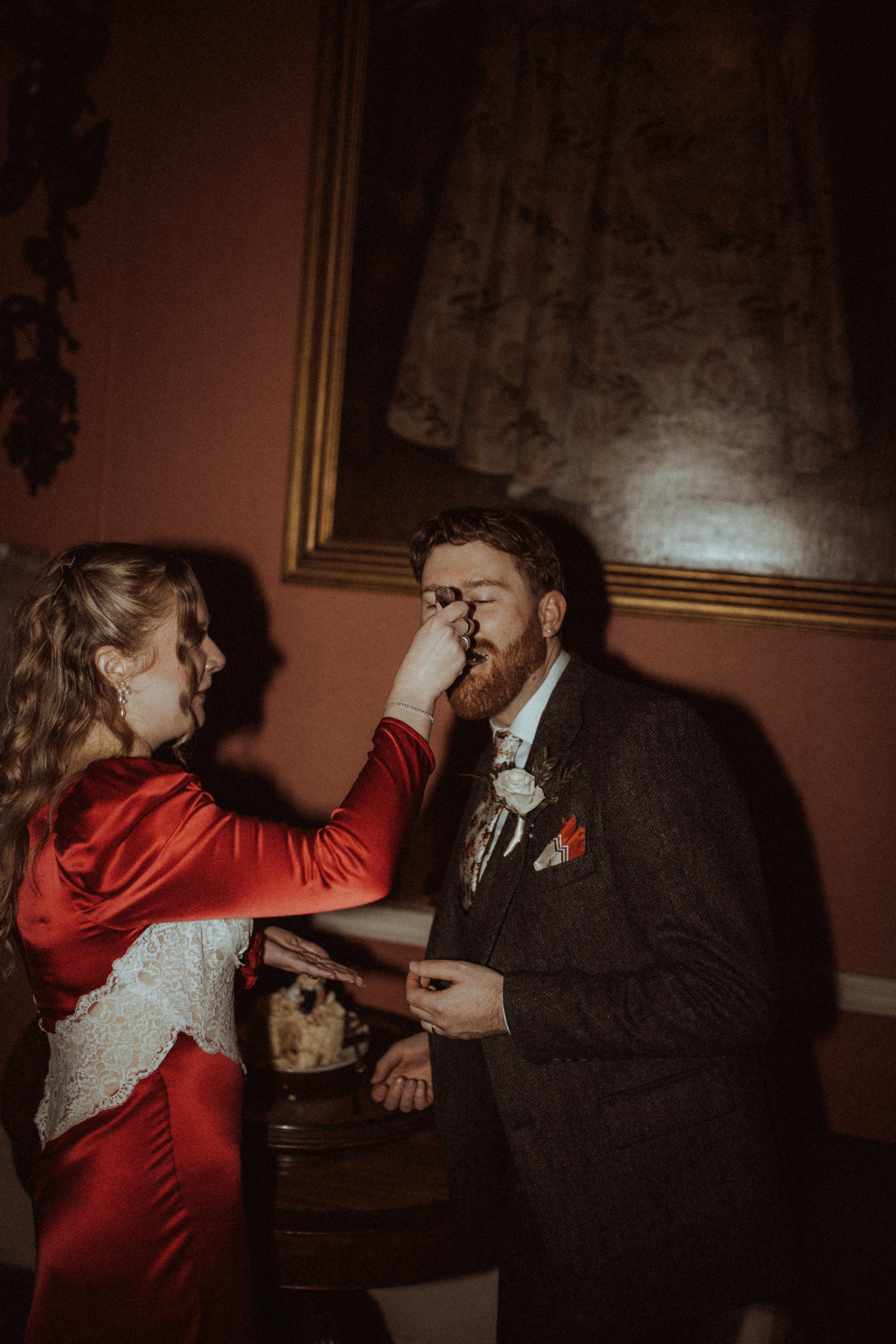 A woman in a red dress is applying makeup to a man in a gray suit at a formal event or wedding.