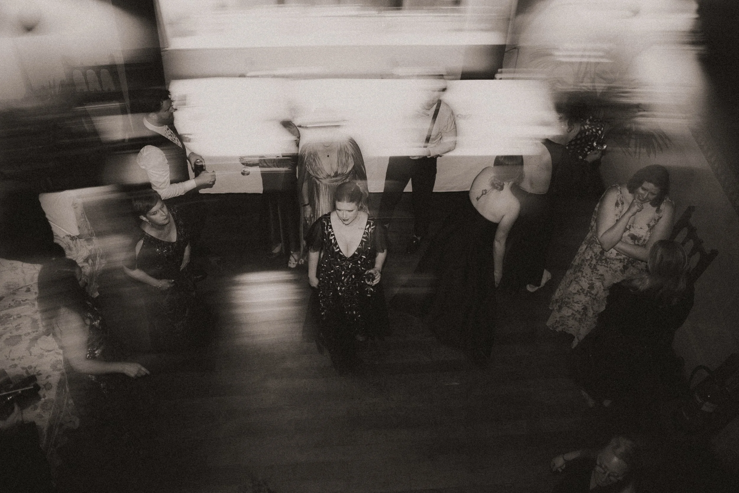 Black and white photo of people at a social gathering viewed from above, with some dancing and others talking or sitting, and motion blur creating a dynamic, lively atmosphere.