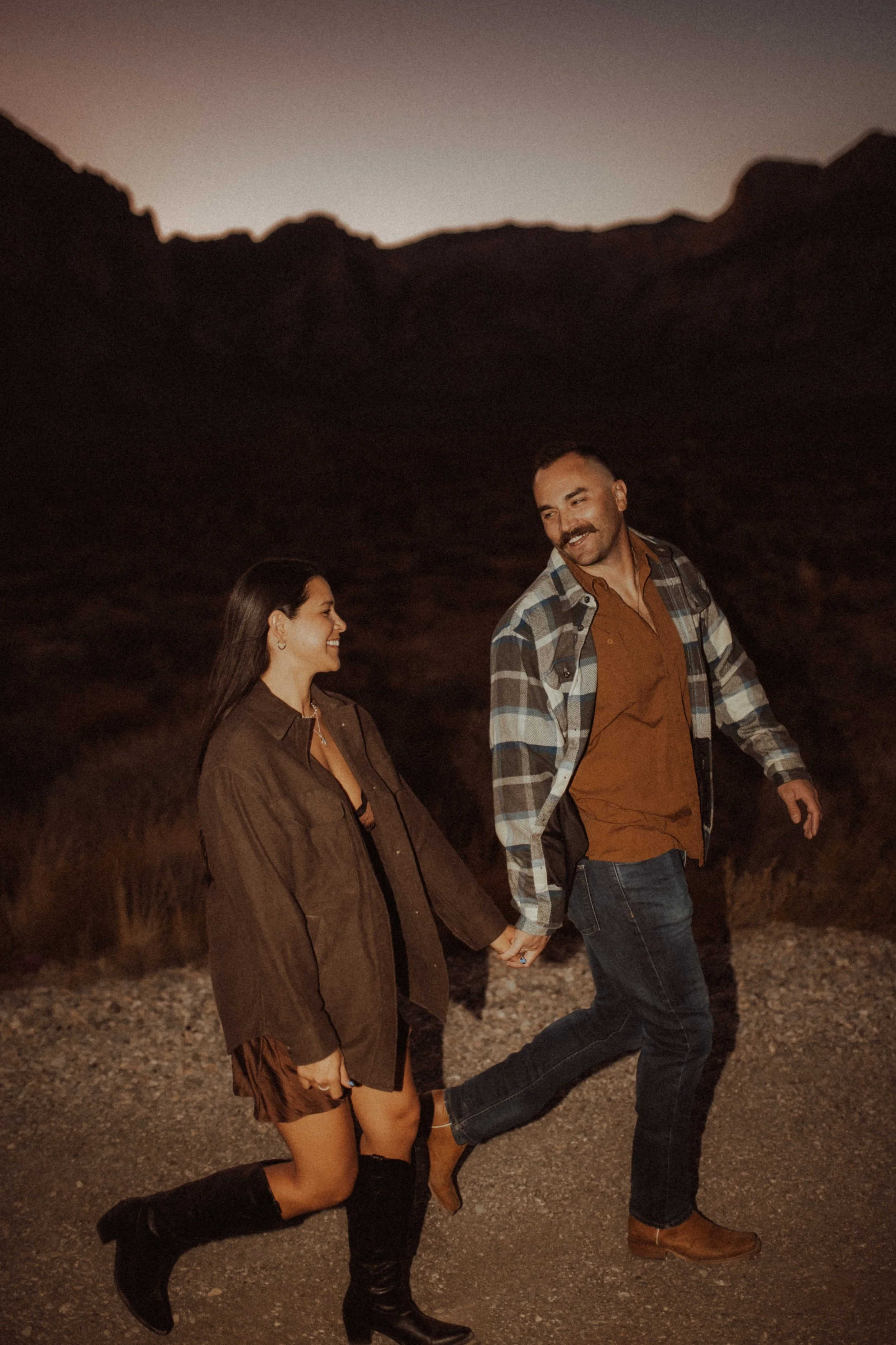 A couple walking hand in hand outdoors during dusk, smiling at each other, with mountain silhouettes in the background.