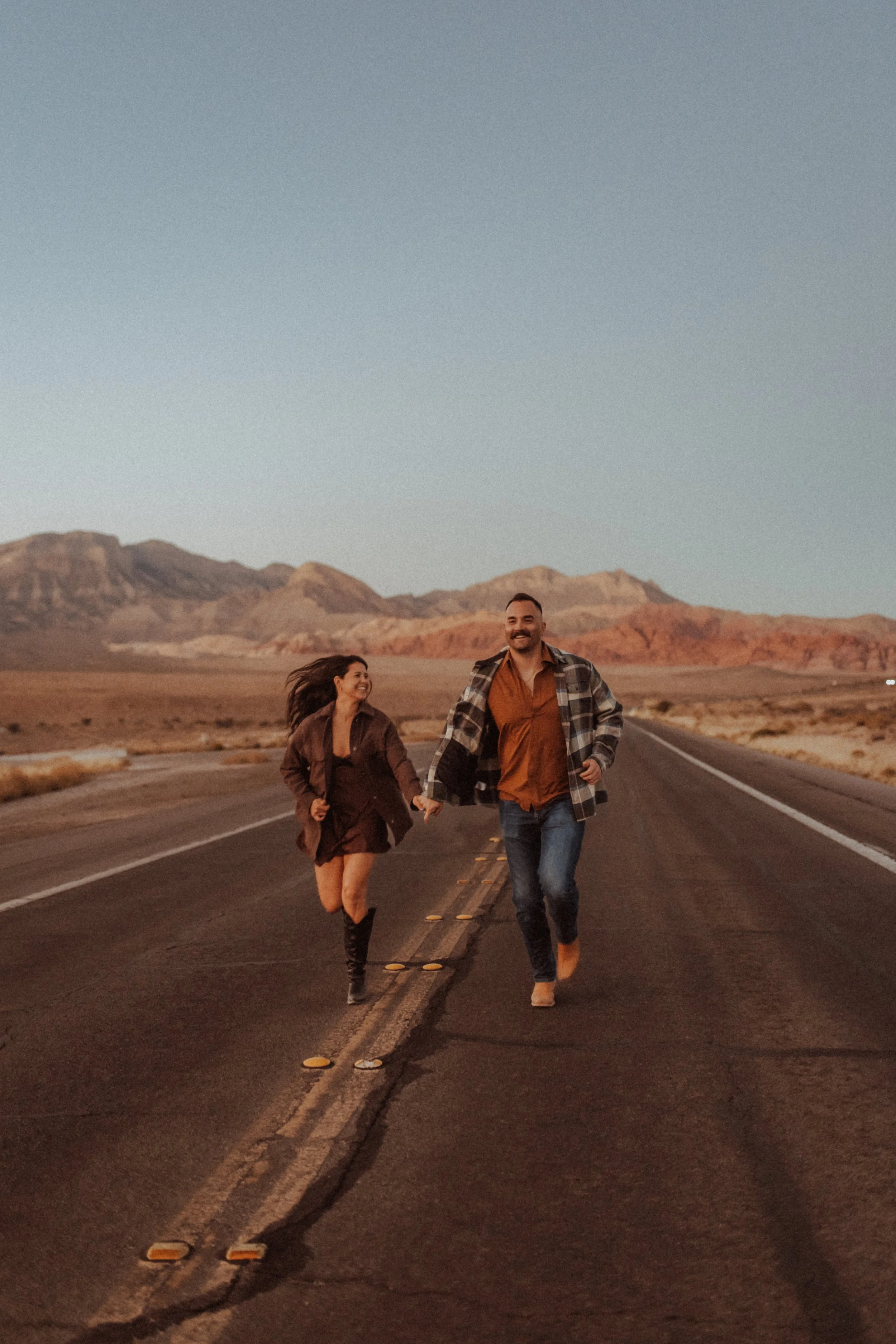 A man and woman holding hands and running down an empty desert road with mountains in the background at sunset.