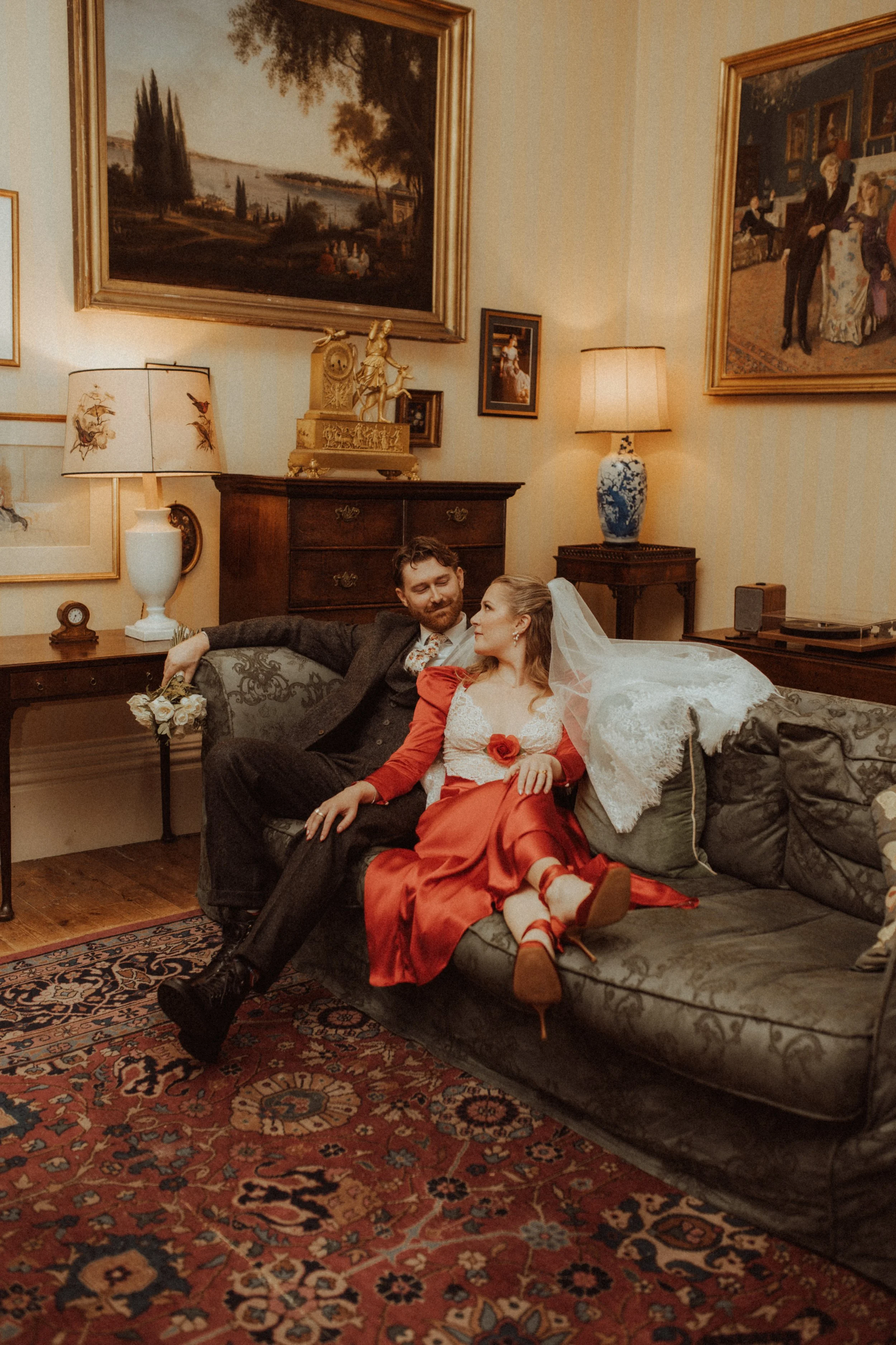 A couple sitting on a vintage-style sofa in a decorated living room, dressed in wedding attire. The woman is wearing a red dress with a white lace top and veil, and the man is wearing a dark suit. The room has classic decor, framed paintings, lamps, and a patterned rug.