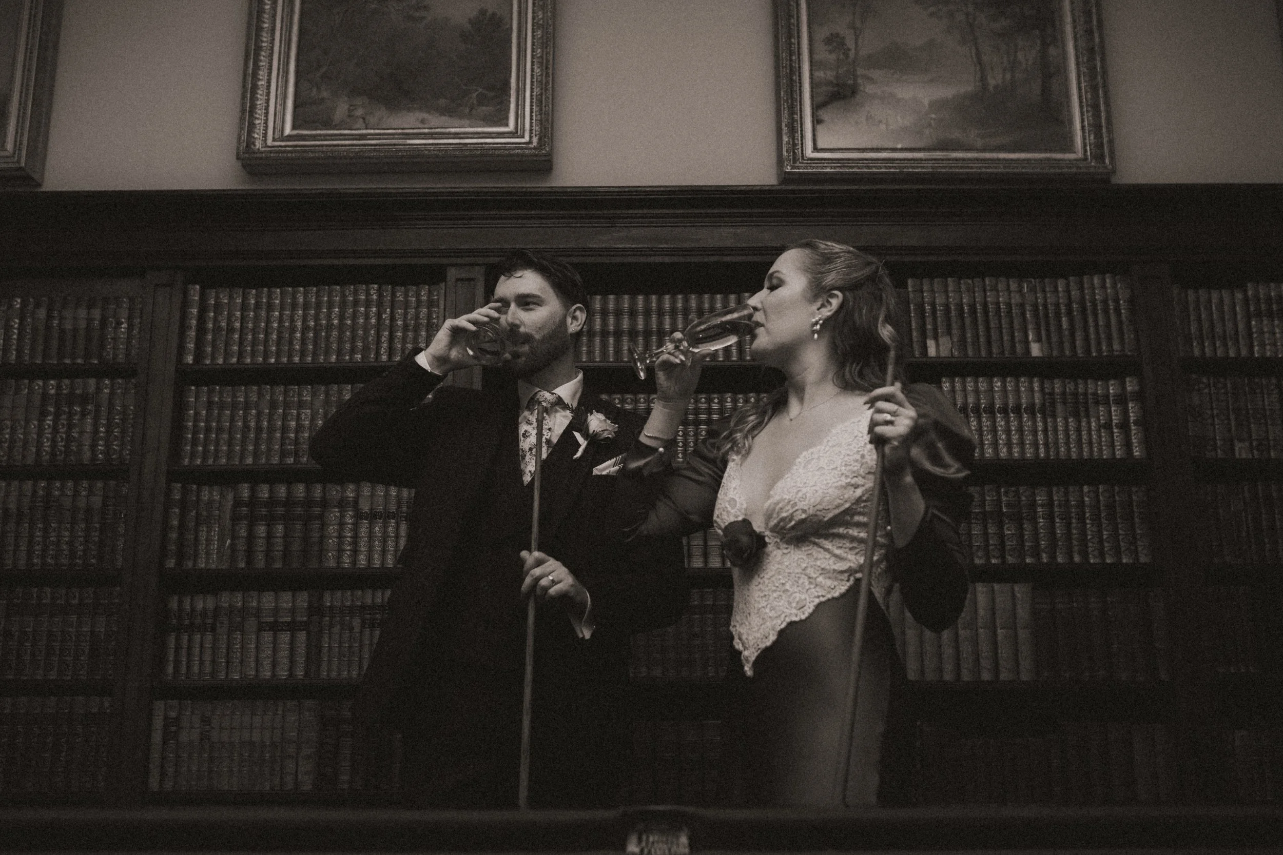 A man and woman dressed in formal attire, holding glasses of wine and toasting, standing in front of a large bookshelf filled with leather-bound books in a dimly lit, elegant room.