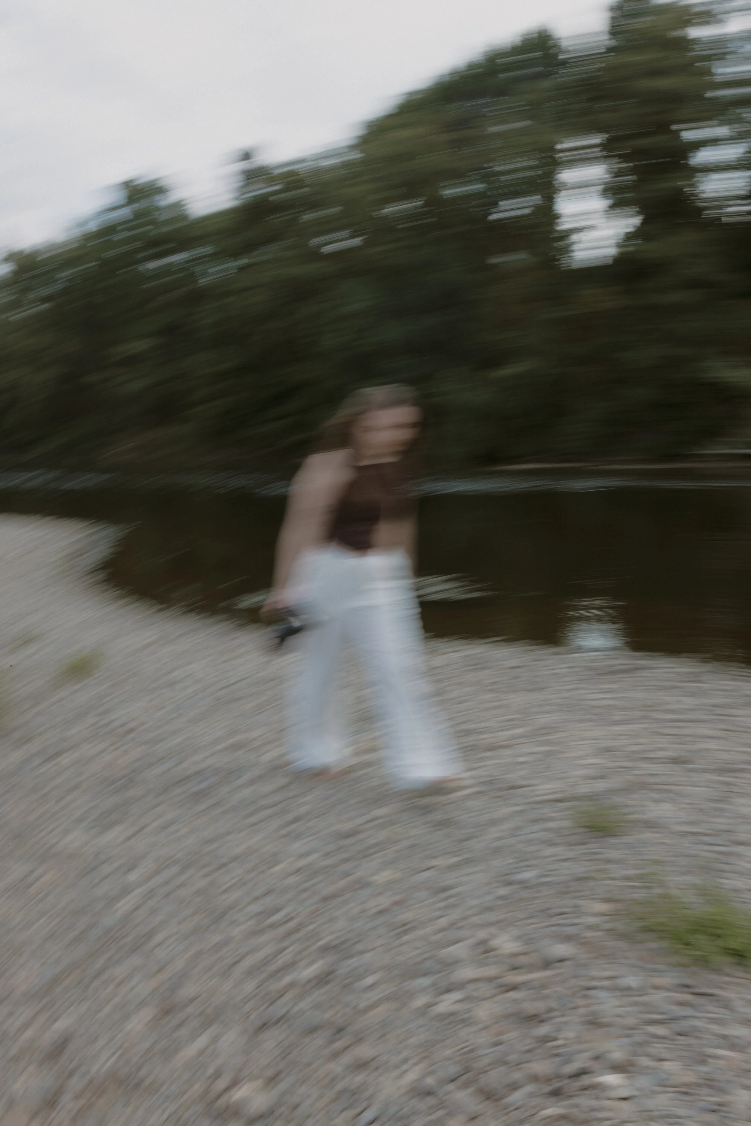 A blurry photo of a woman with long brown hair in a black sleeveless top and white pants, standing on a rocky shoreline near a body of water with trees in the background.