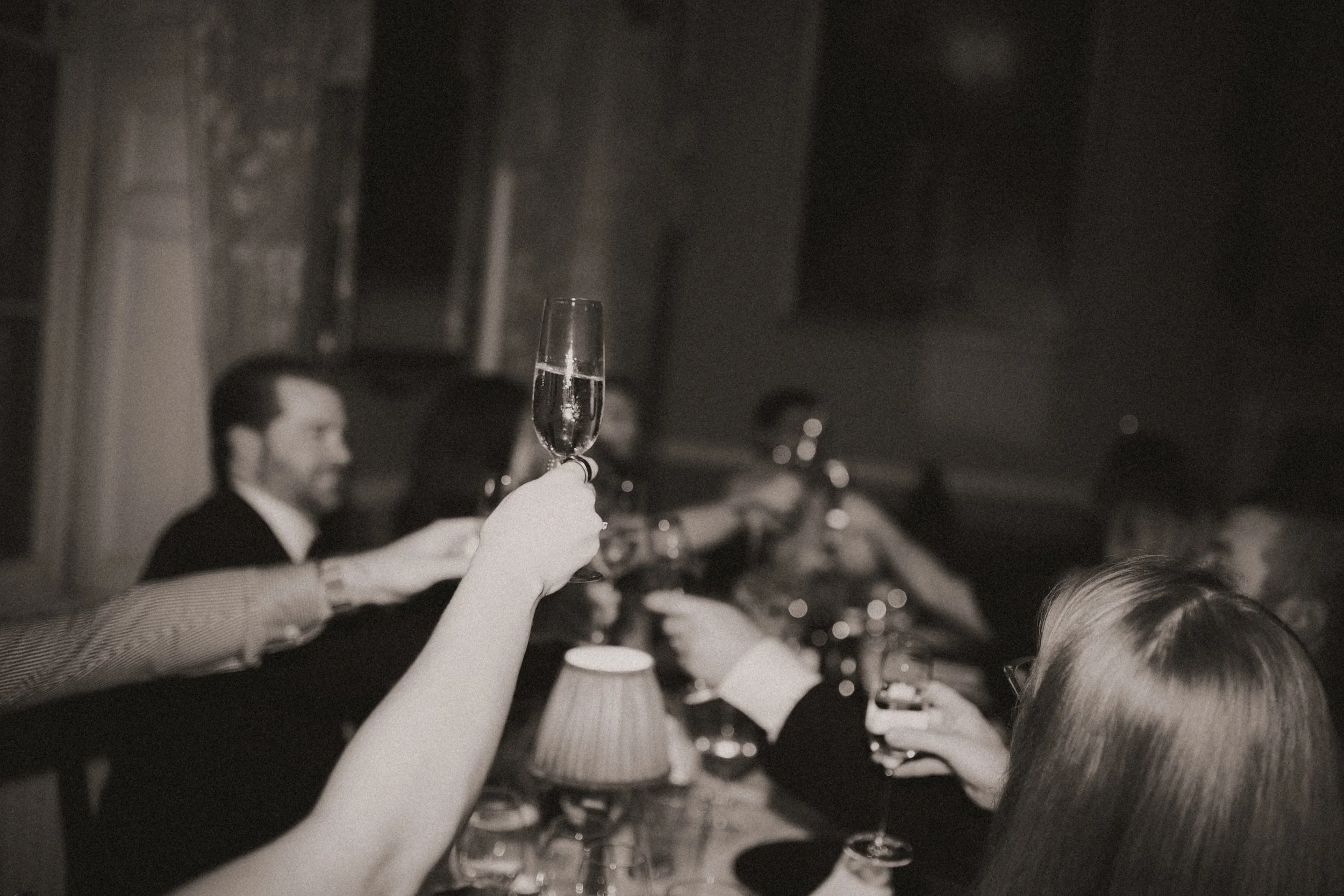 People at a dimly lit dinner party raising glasses in a toast, including a woman holding a champagne flute in the foreground.