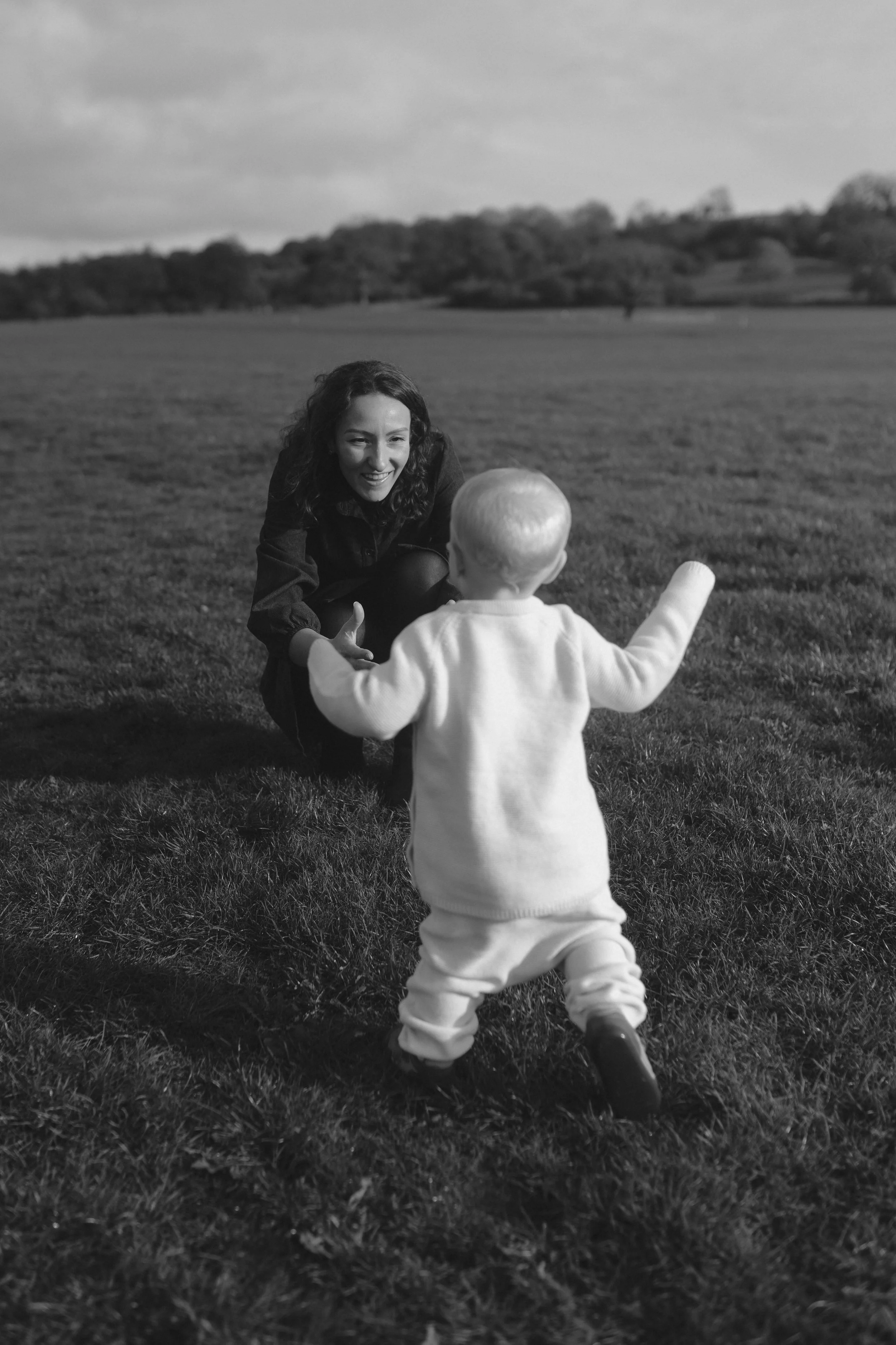 A woman crouching in a grassy field, smiling and reaching out to a toddler who is walking towards her with arms open.