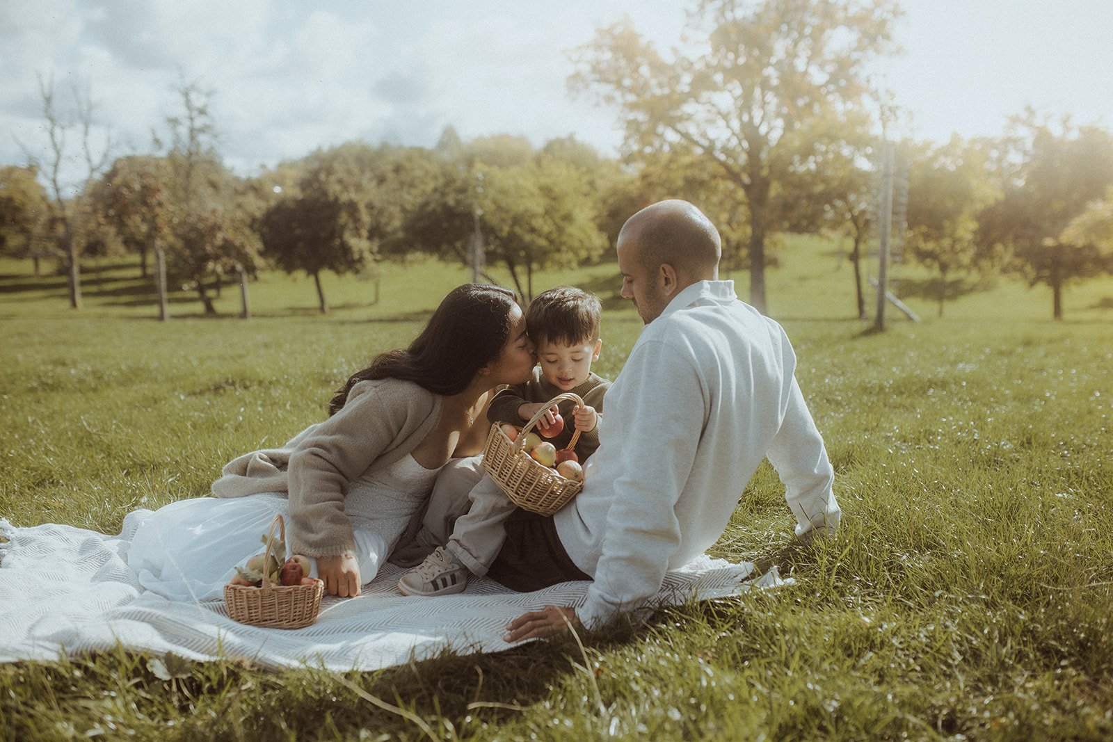 A family of three with a woman, a man, and a young boy having a picnic on a grassy field in a park during daytime.
