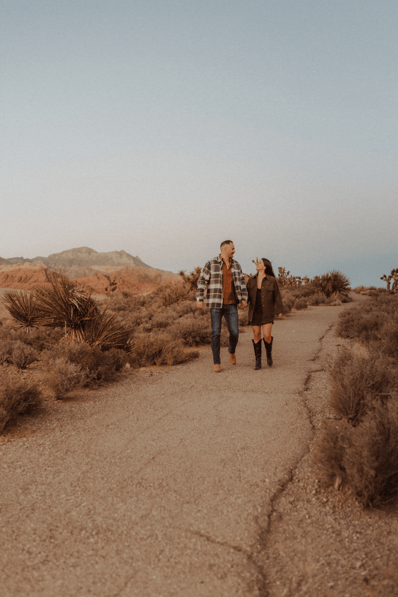 A couple walking hand in hand on a desert trail at sunset, surrounded by desert plants and mountains in the background.