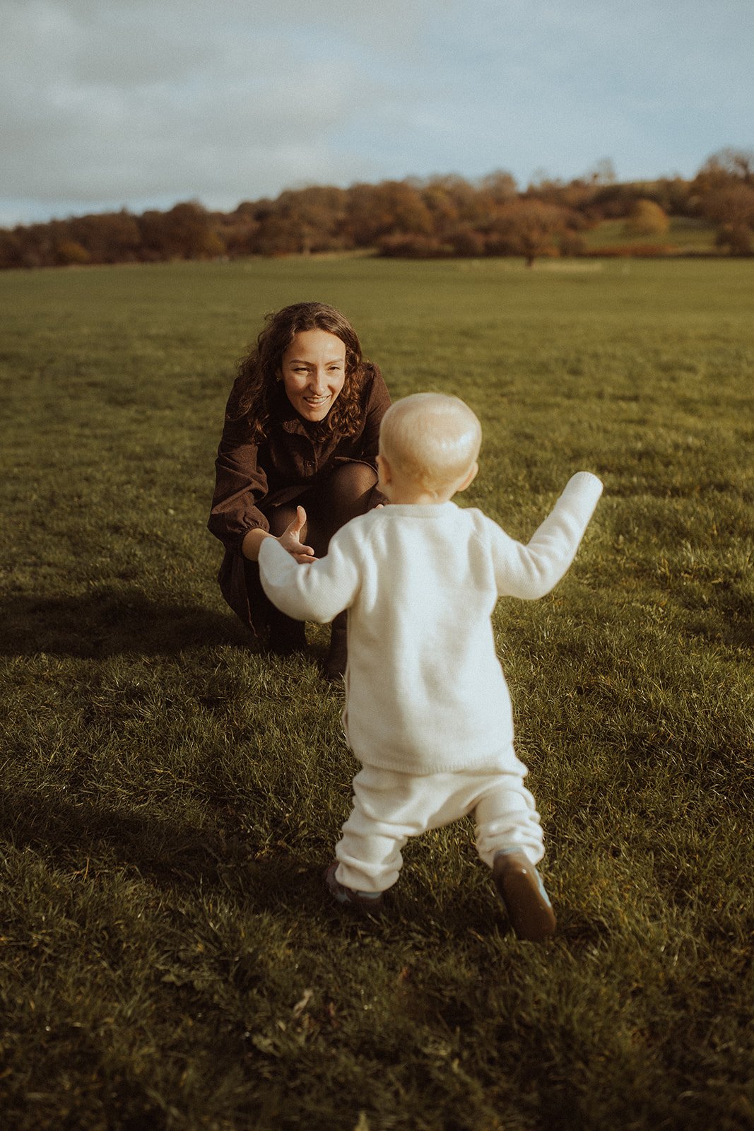 A woman smiling and reaching out to a toddler who is walking towards her on a grassy field, with trees and a cloudy sky in the background.