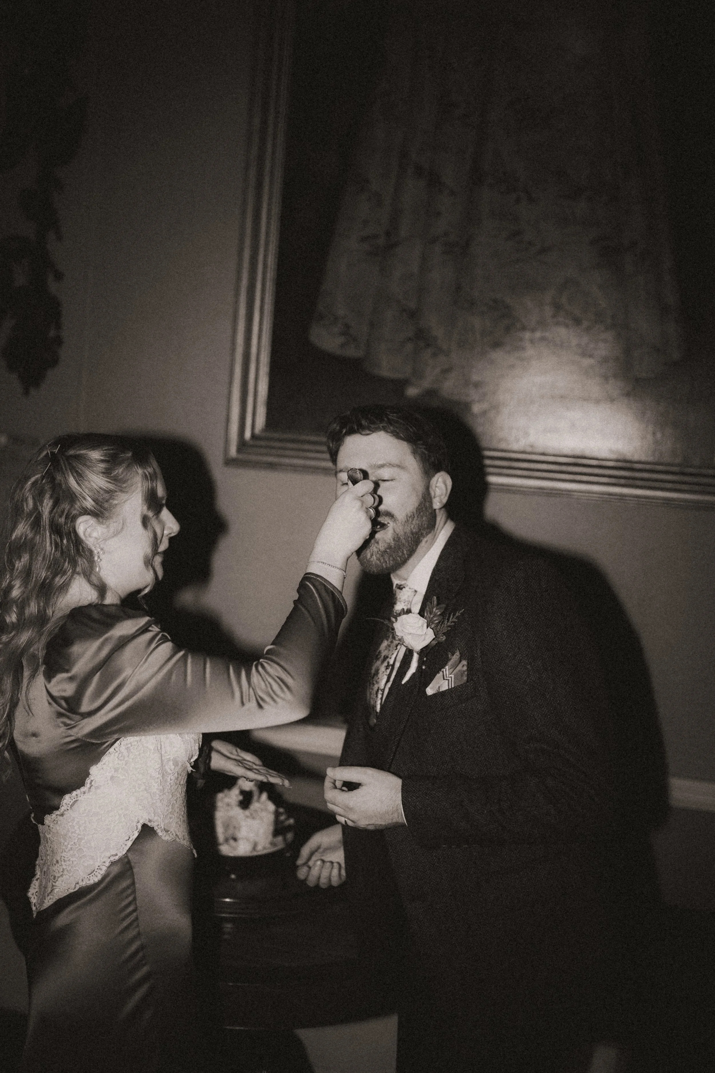 A woman applying makeup or face paint to a man dressed in formal attire at a wedding or formal event, indoors with a large framed painting on the wall behind them.