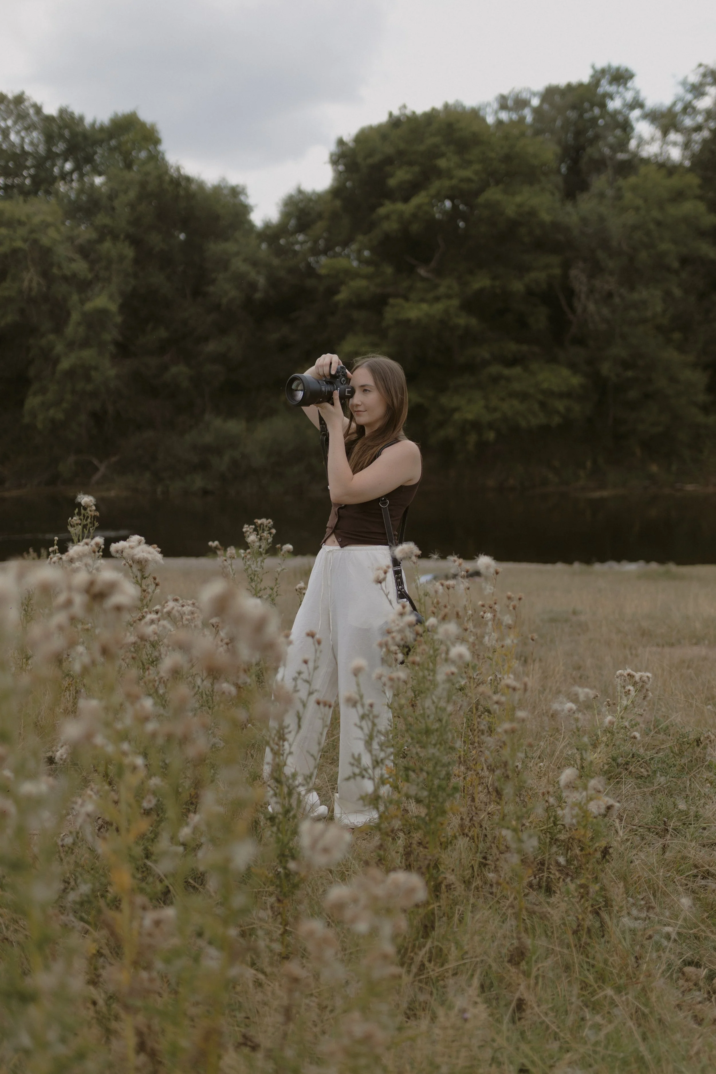 A woman standing in a field with wildflowers, holding a camera up to take a photo, with trees and a river in the background.