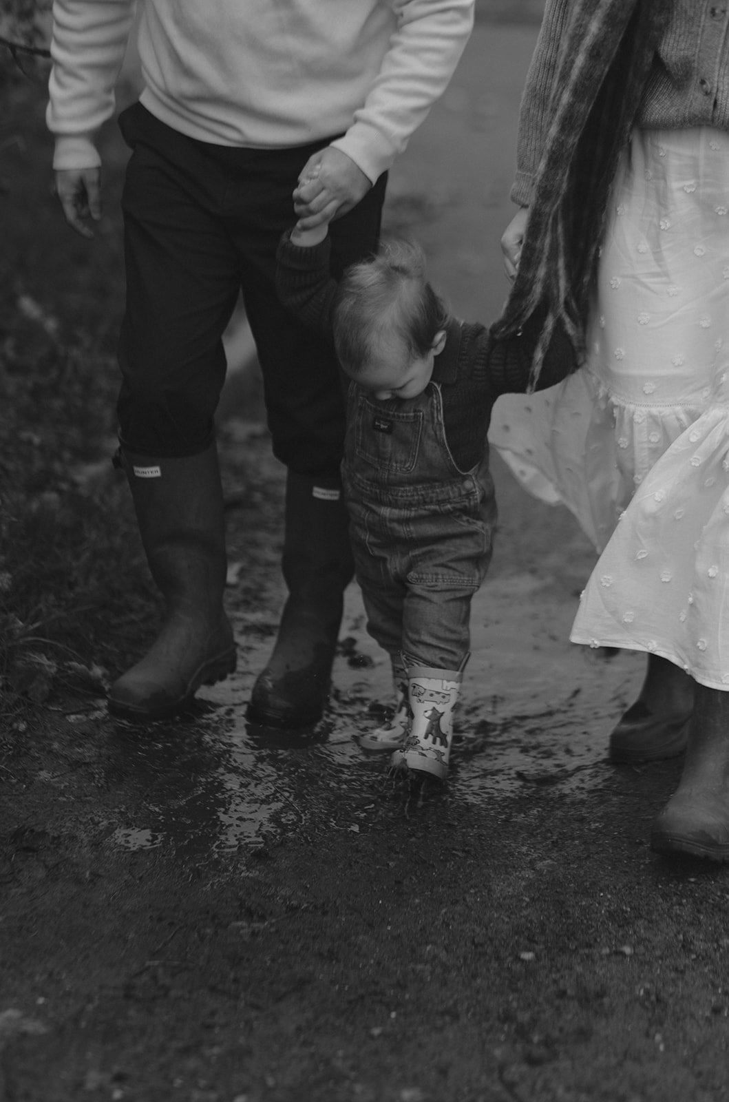 A young boy walking through a puddle with help from two adults, one holding his hand and one guiding him, on a muddy outdoor trail.