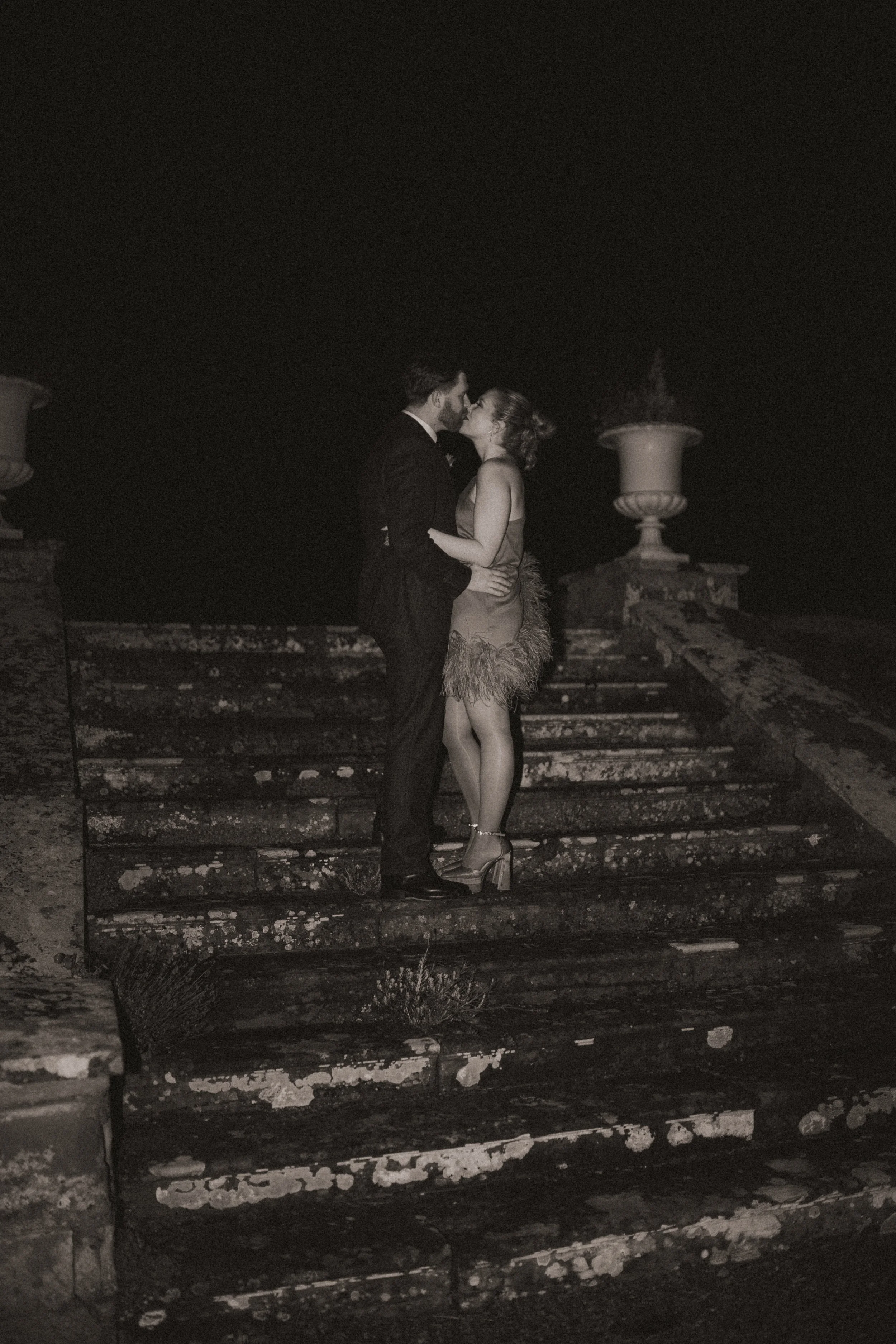A black and white photo of a couple kissing on a set of old, weathered stairs at night. The man is dressed in a suit, and the woman is wearing a dress with feather details and high heels. There are large flower pots on either side of the stairs.