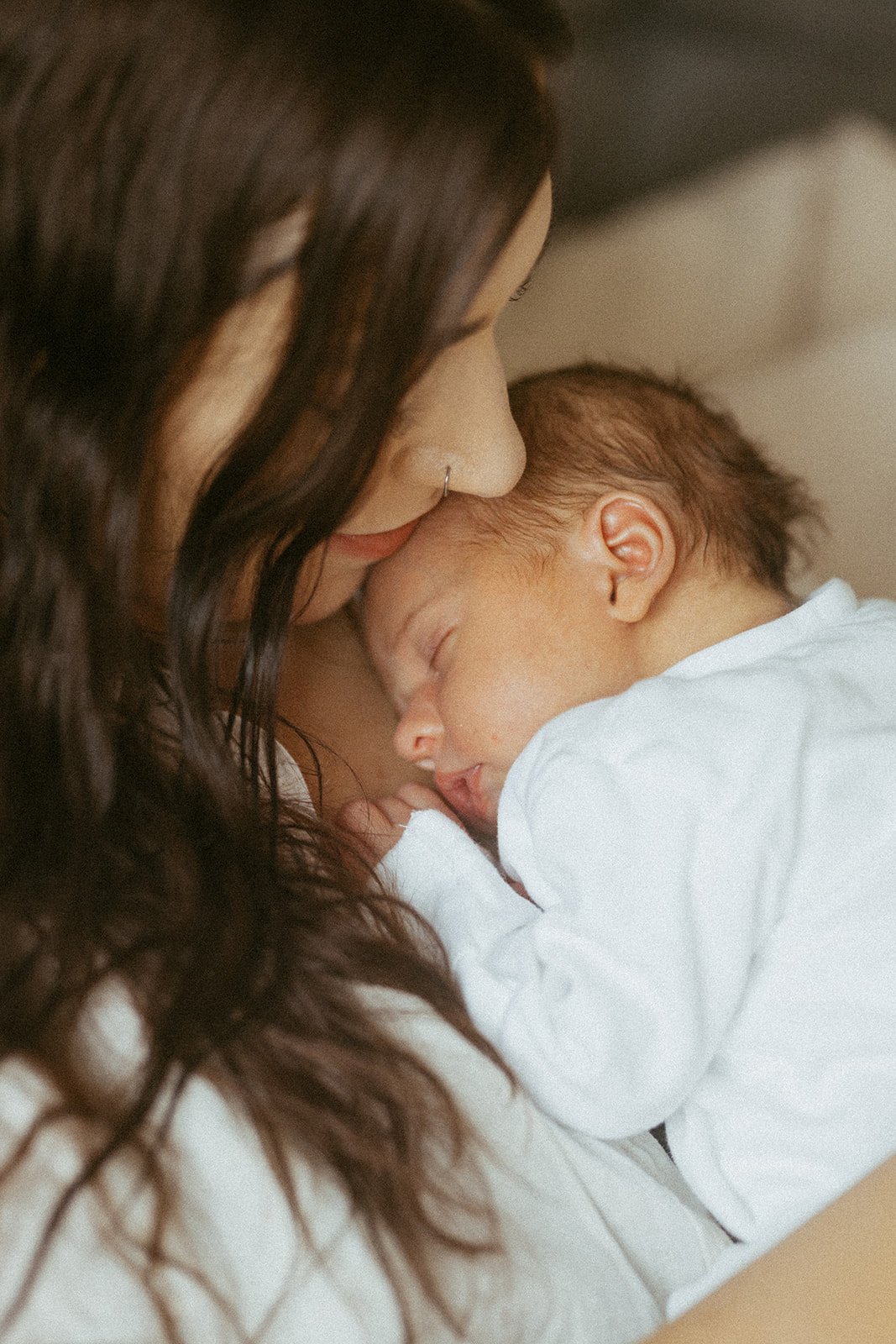 A woman with long dark hair and a nose ring tenderly holds and kisses a sleeping child's forehead, both dressed in white.
