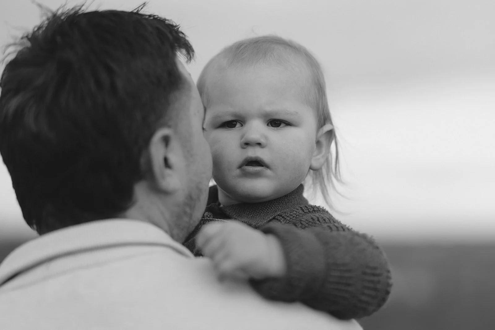 A young child with light hair being held by an adult man with dark hair. The child appears to be looking at the man with a serious expression.