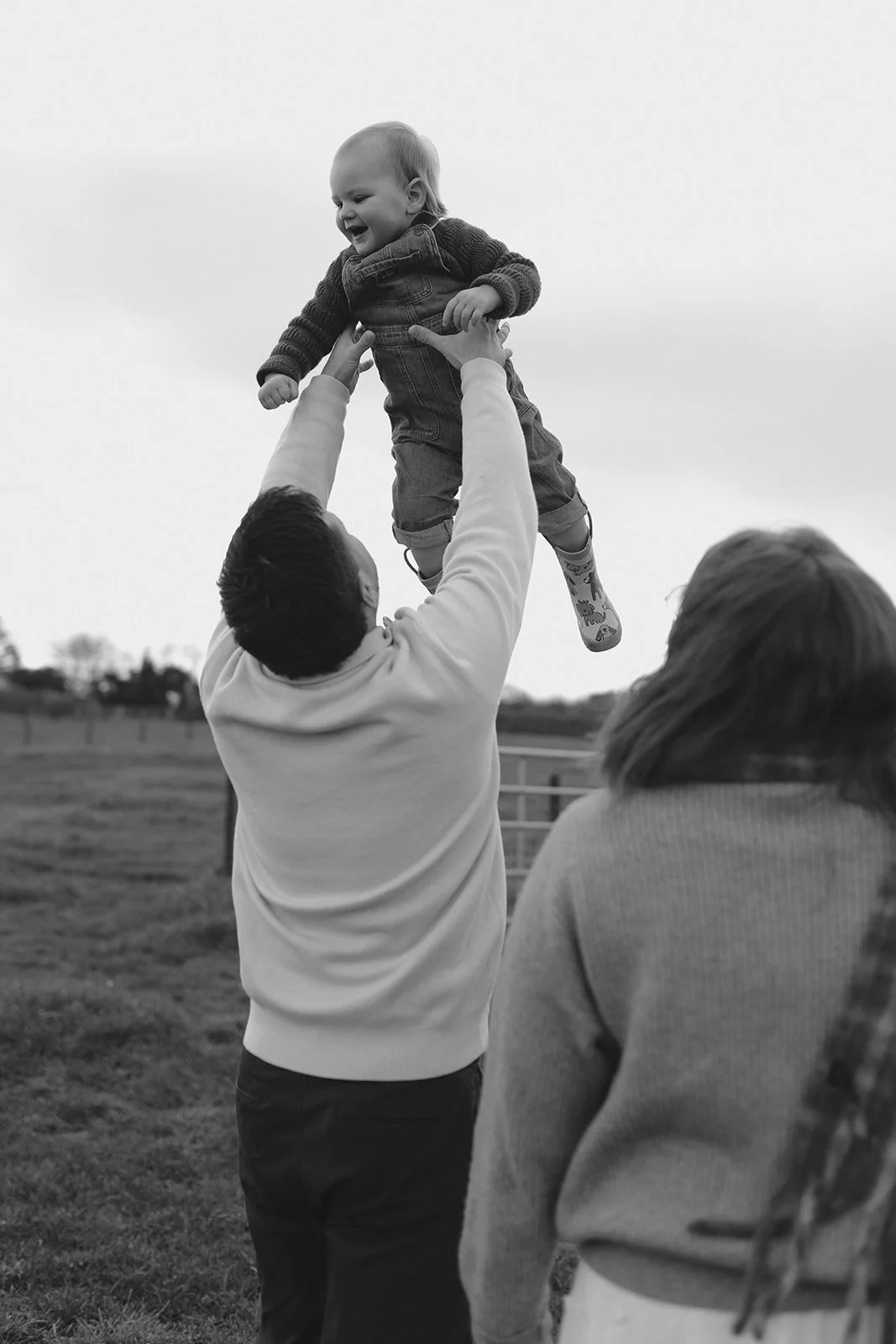 A man lifting a laughing child into the air outdoors, with two women watching nearby.