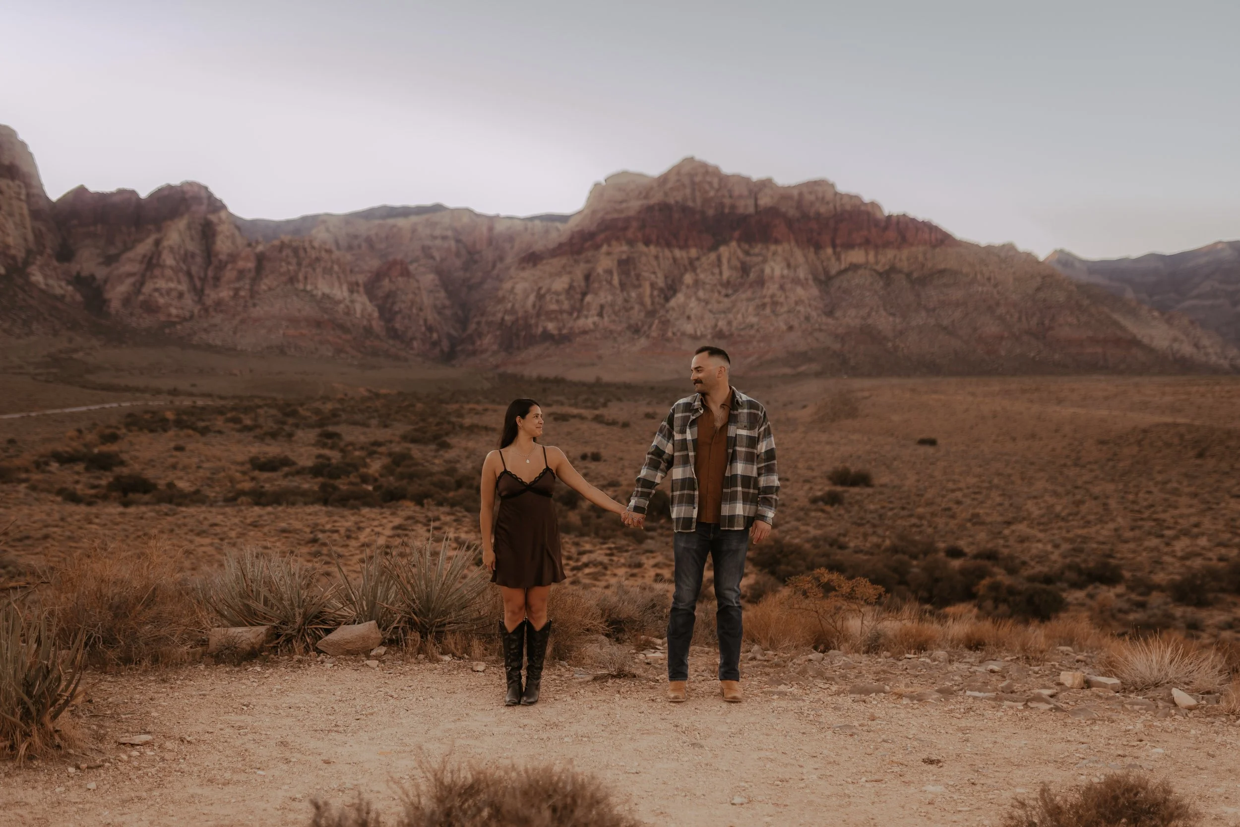 A couple holding hands walks in a desert landscape with mountains in the background during sunset.