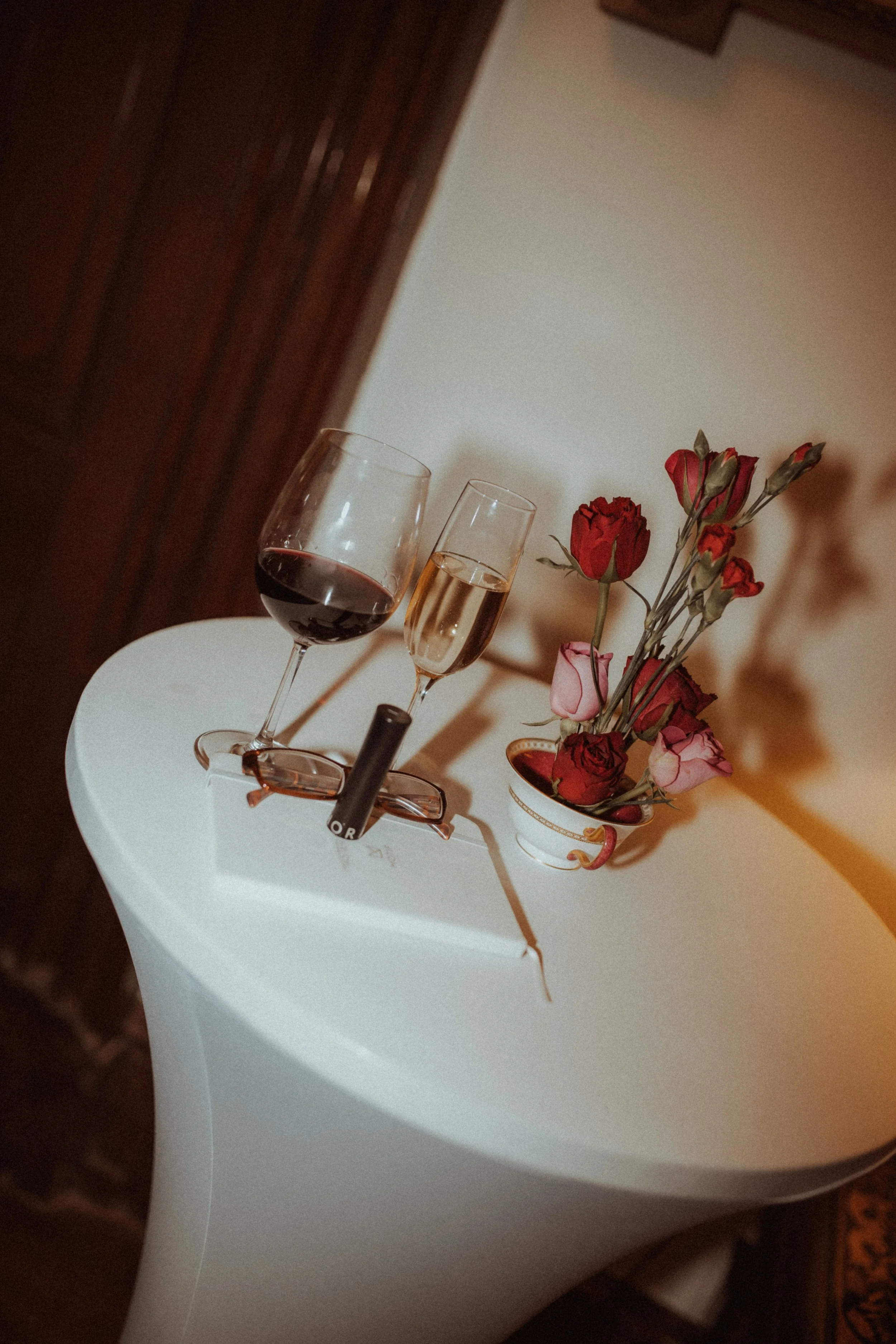 A white table with a pair of glasses, a wine glass filled with red wine, a champagne flute filled with champagne, a small decorative vase with red and pink roses, and a pair of glasses.