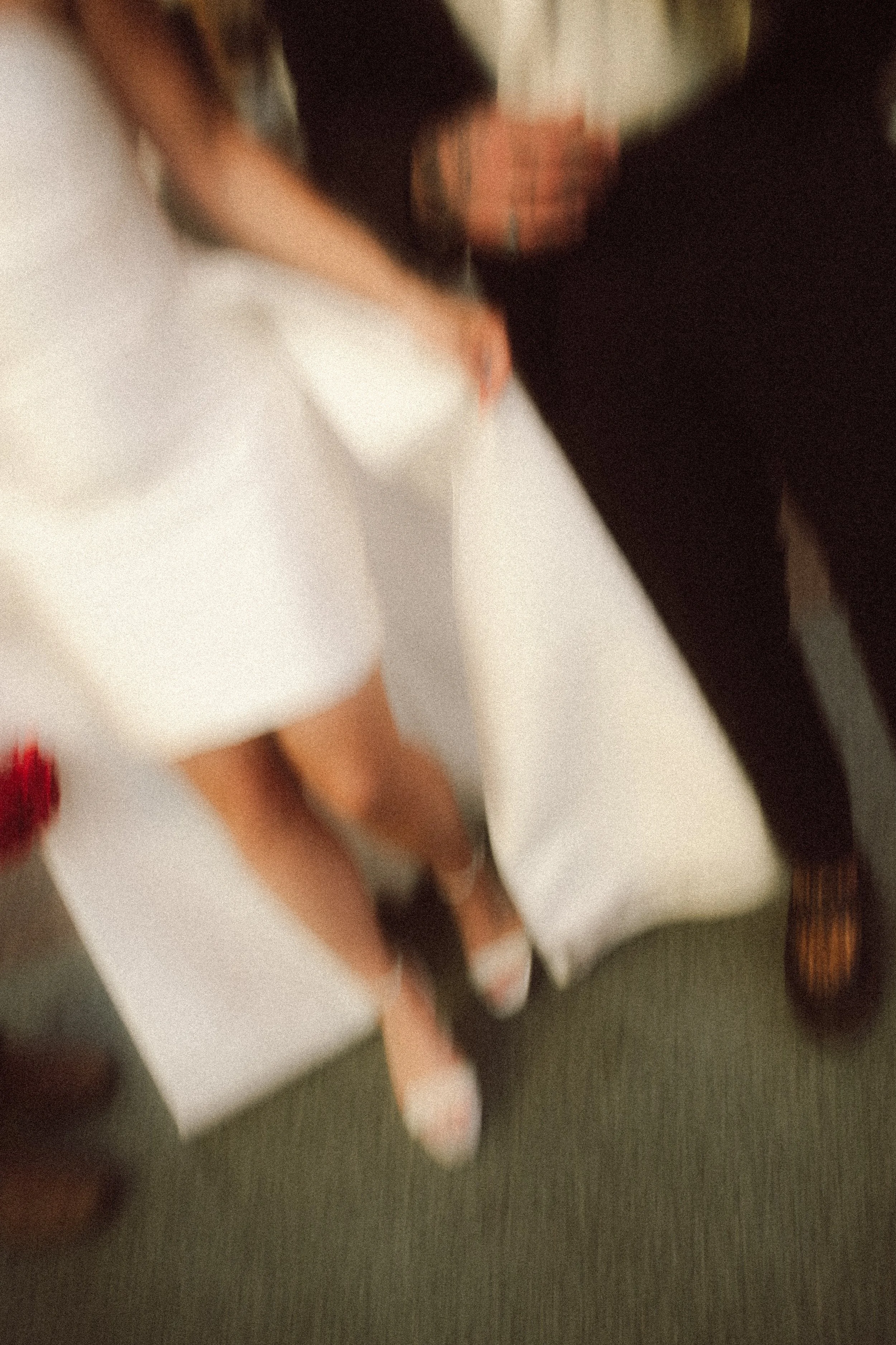 Blurred photo showing a woman in a white dress and heels, standing near a bed, with a person in black pants and shoes standing beside her.