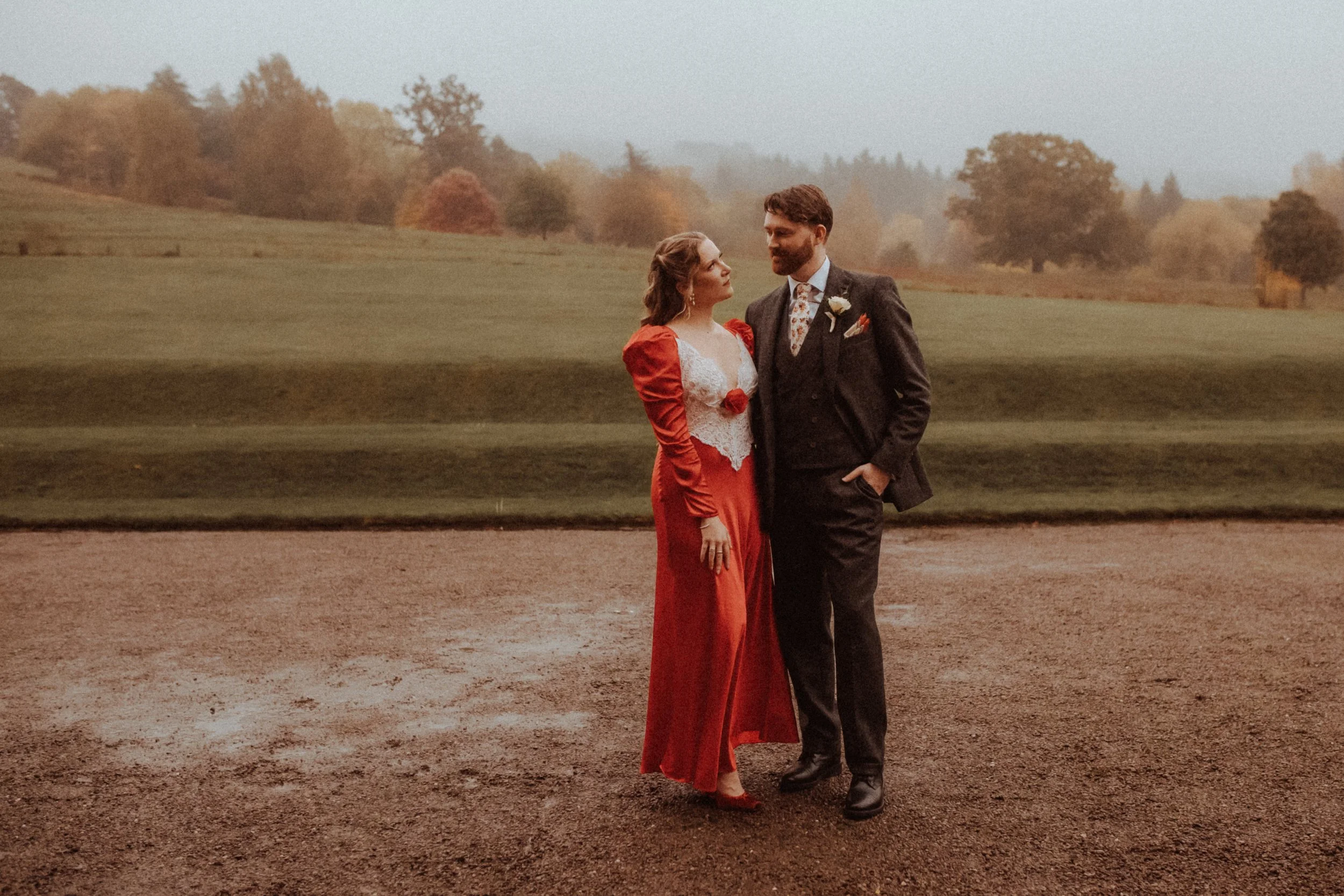 A couple dressed in vintage wedding attire standing on a dirt field with an open grassy area and trees in the background. The woman wears a red dress with a white lace bodice and matching red heels, and the man wears a dark suit with a floral vest and a boutonniere.