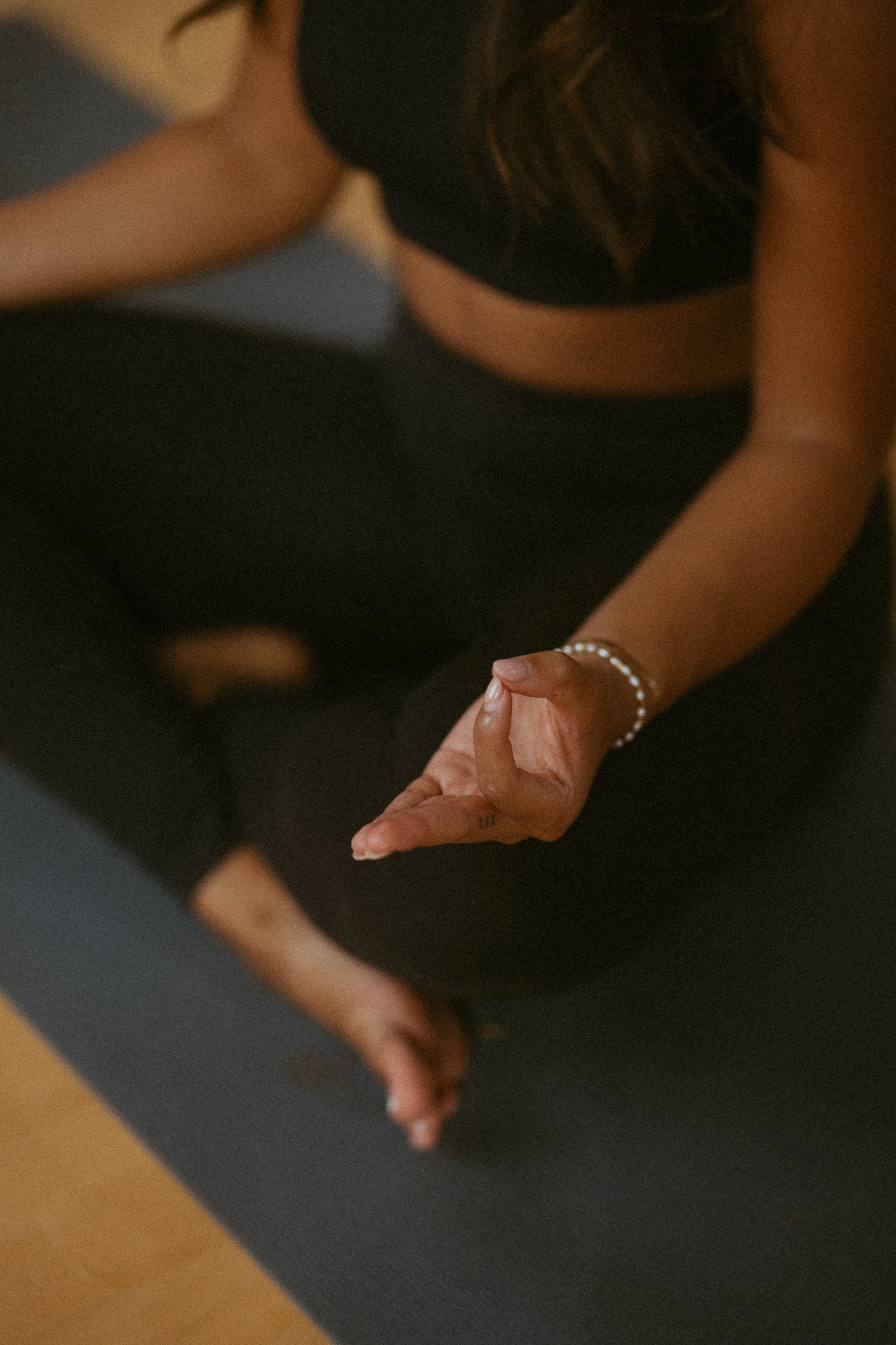 Close-up of a woman in a cross-legged yoga pose with her hands in a mudra position, wearing black clothing and a pearl bracelet, on a yoga mat.
