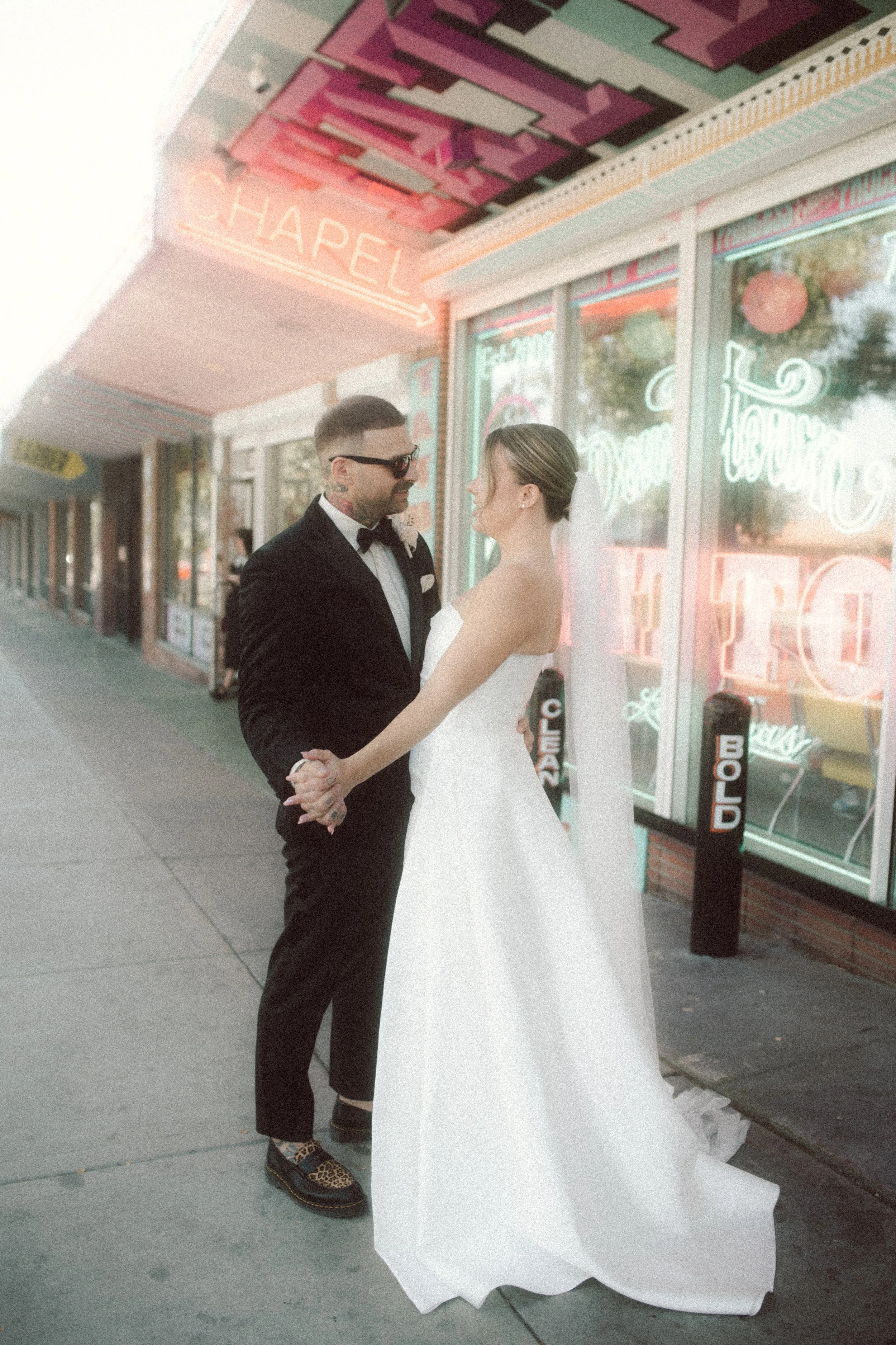 A bride and groom stand outside a neon-lit shop on a city sidewalk, holding hands and gazing at each other. The bride wears a white strapless wedding gown with a veil, and the groom is in a black tuxedo with glasses. Neon signs and colorful storefront windows illuminate the scene.