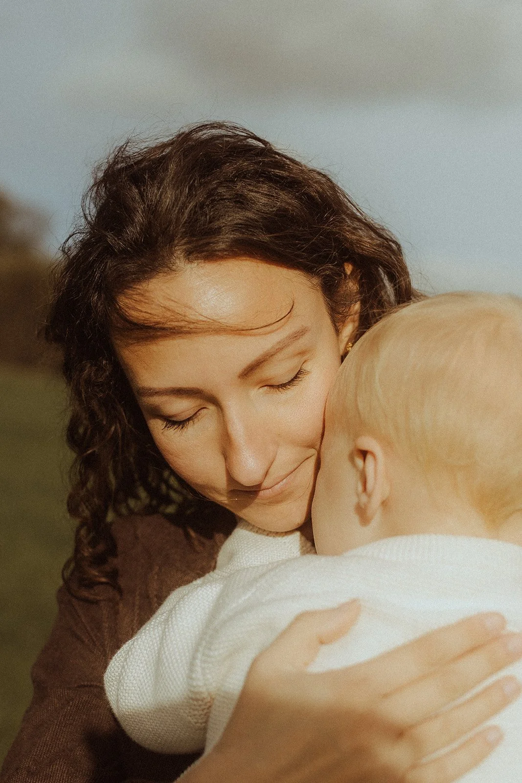 Close-up of a woman with curly hair smiling gently as she holds a young blonde-haired child close to her, both with eyes closed, outdoors during daytime.