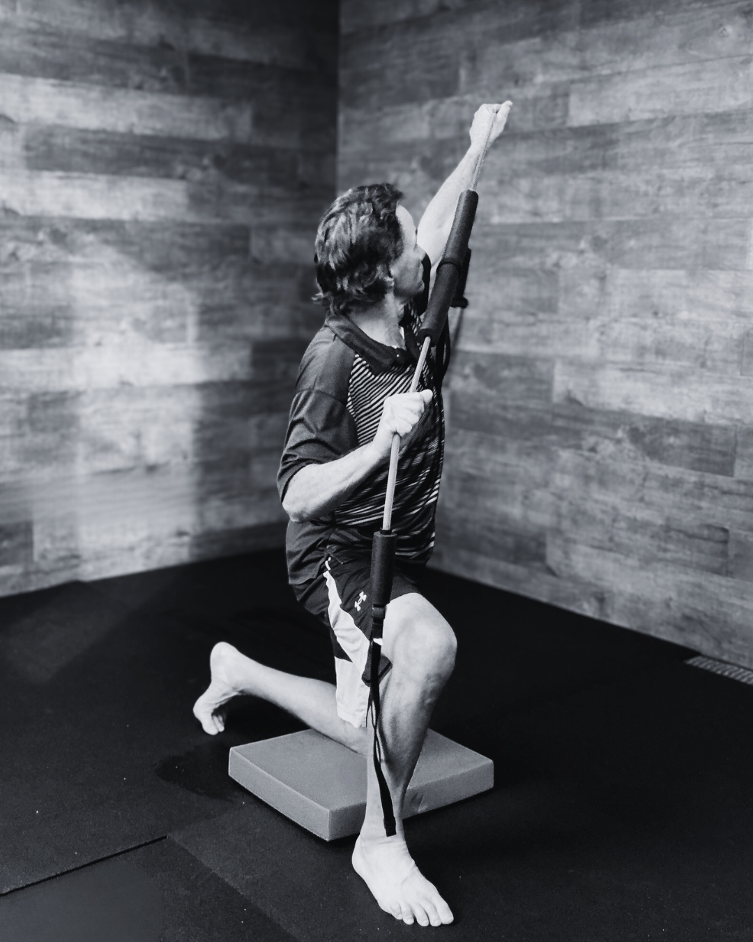 A person performing a stretch exercise using a resistance band while kneeling on a cushion in a room with wood-paneled walls.