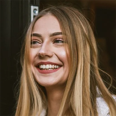 A young woman with long blonde hair smiling in front of a dark background.