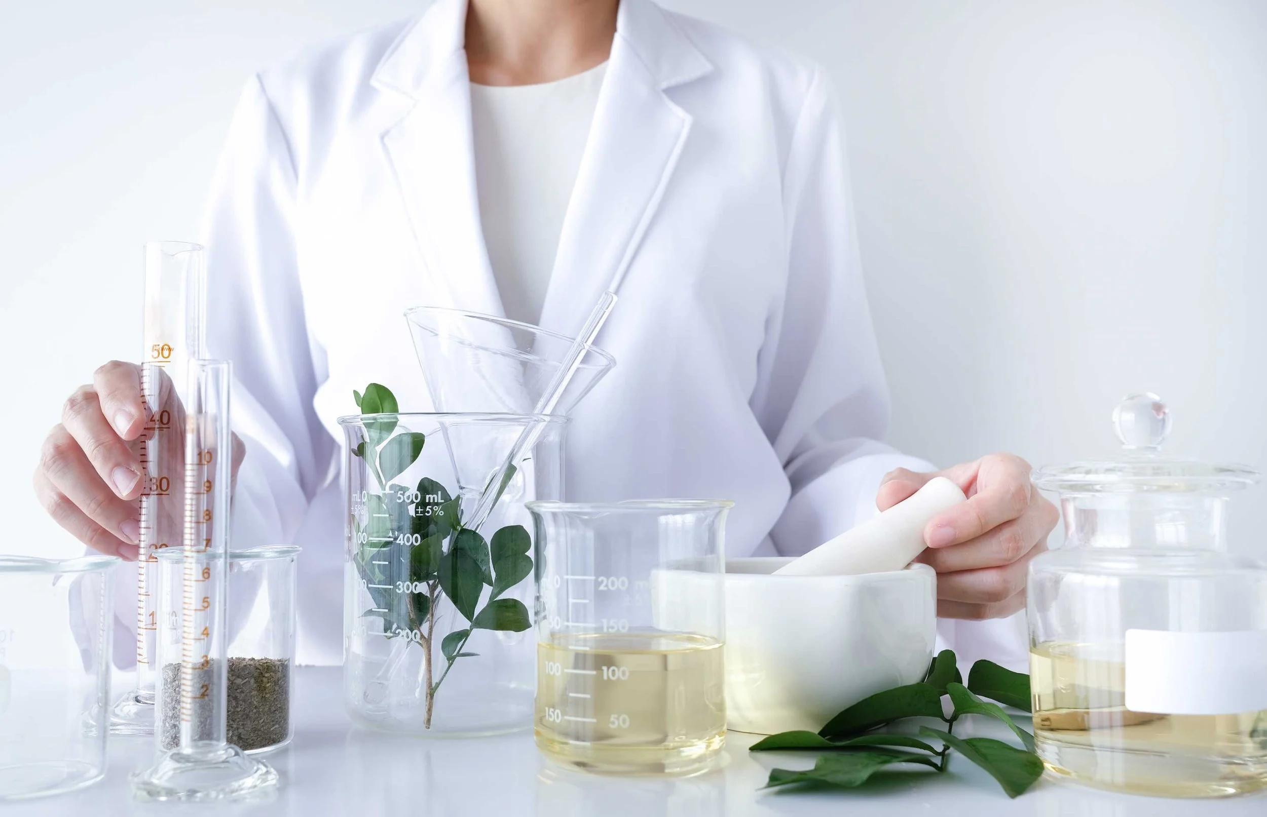 Scientist in white lab coat working with glass beakers and test tubes containing liquids and plant samples in a laboratory.
