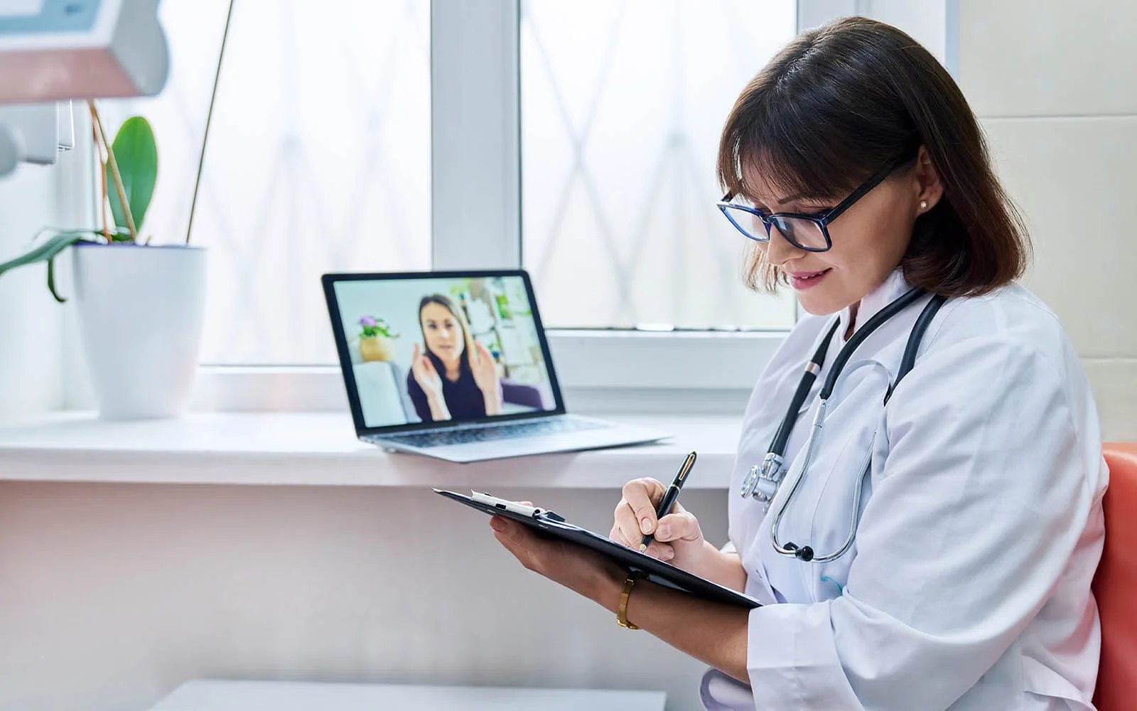 A female doctor with glasses, wearing a white coat with a stethoscope around her neck, writing on a clipboard at her desk. In the background on a laptop screen, a woman is making a gesture with her hands during a video call.