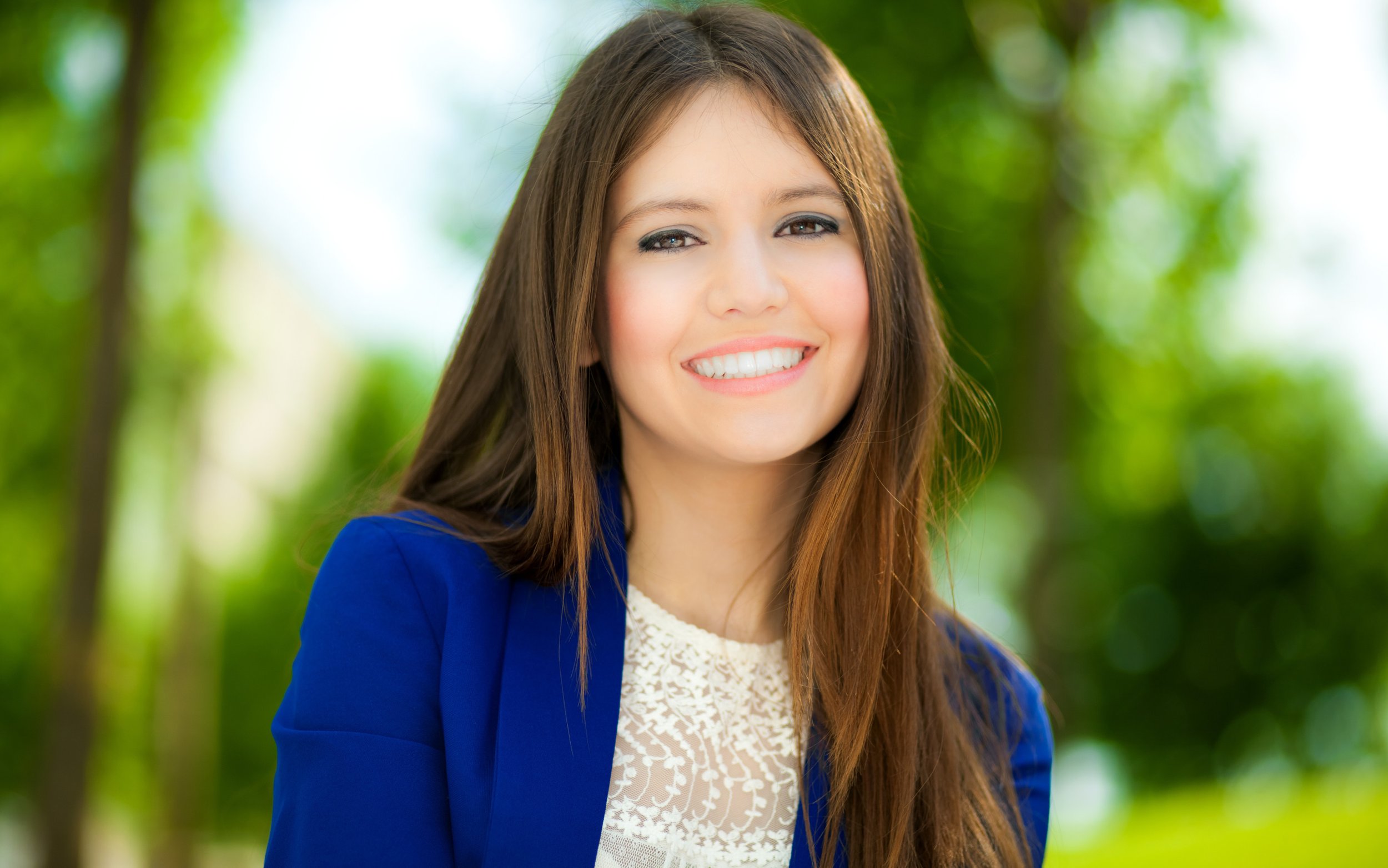 A young woman with long brown hair, wearing a blue blazer and a white lace top, smiling outdoors with blurred green foliage in the background.