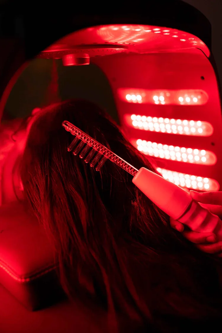 A person with long dark hair undergoing a red light therapy treatment, using a handheld comb device in a red-lit environment.
