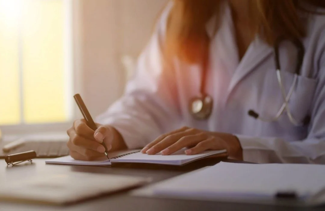 A person in medical attire writing in a notebook at a desk with sunlight coming in through a window.