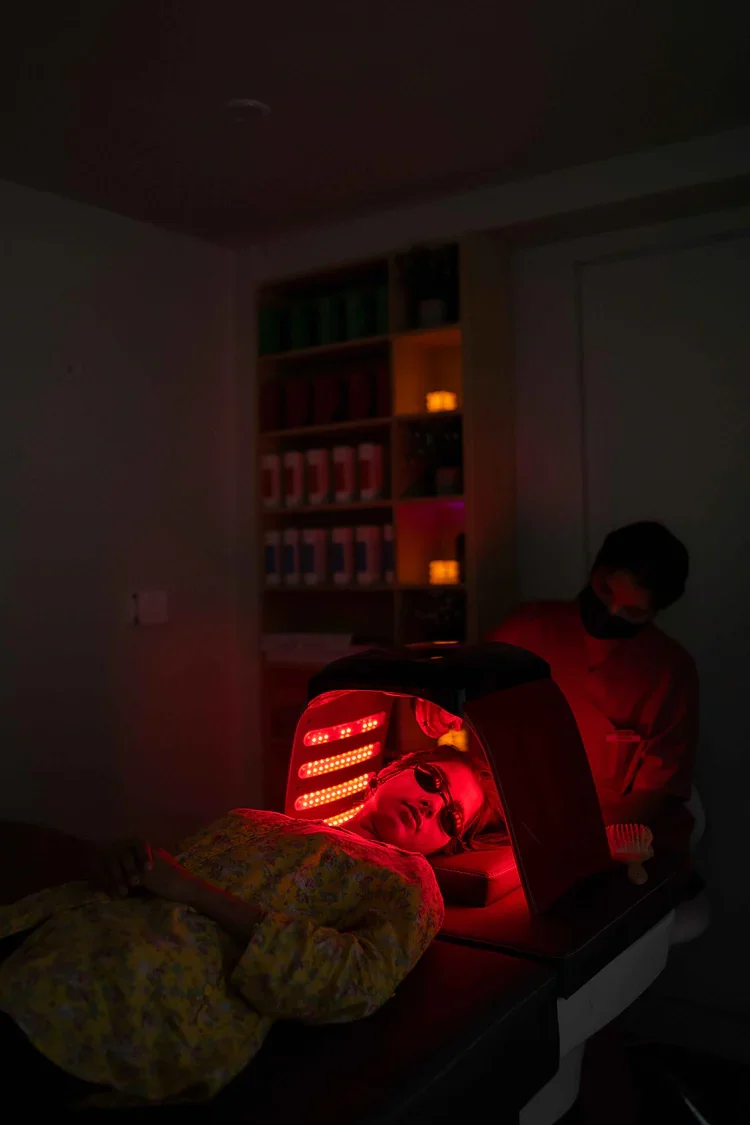 Person lying down under red light inside a machine, with another person standing nearby in a dimly lit room.