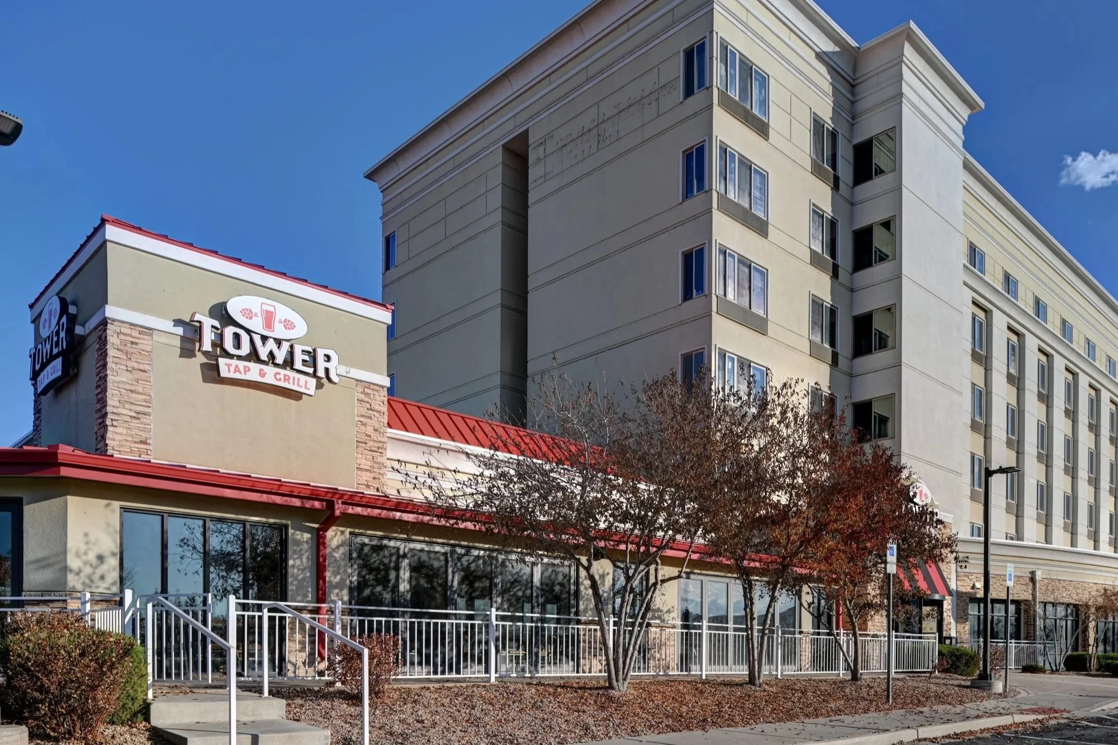 Exterior view of a Tower Tap & Grill restaurant with a large hotel or apartment building in the background. The restaurant has large windows, a red metal roof, and a white fence along the sidewalk. There are trees with autumn leaves and a clear blue 