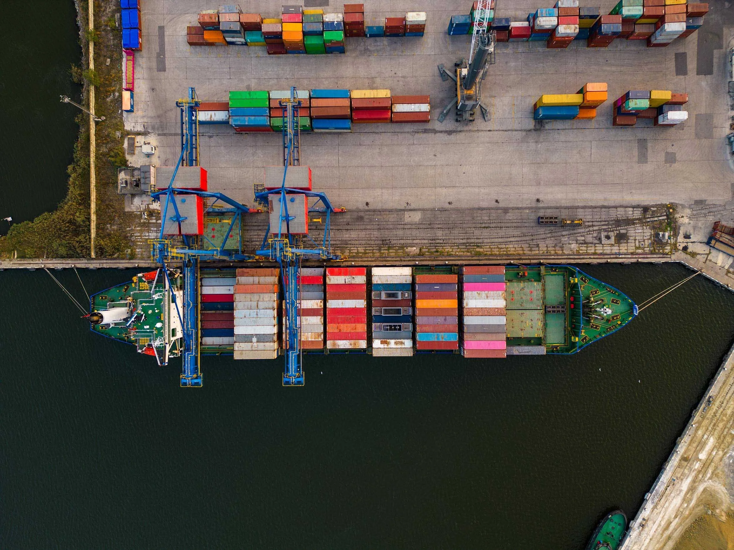 An aerial view of a cargo ship docked at a port with containers on board and on the dock, along with cranes and a waterway.