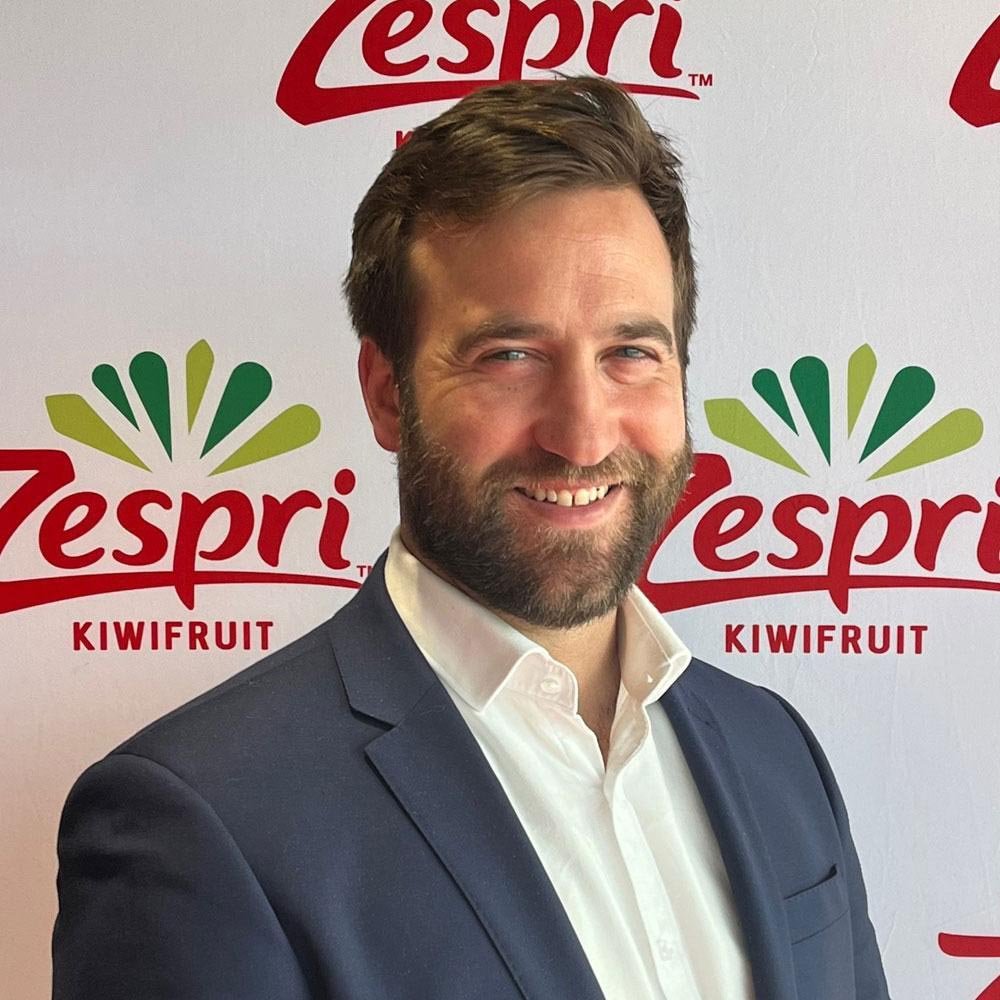 A man with brown hair and a beard, wearing a dark suit and white shirt, smiling at the camera. Behind him is a white backdrop with red and green text and logos for 'Zespri' and 'Kiwifruit.'