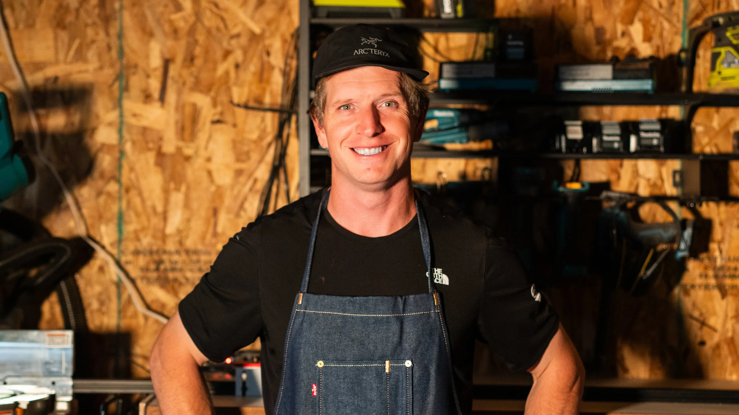A smiling man wearing a black cap and a dark shirt with an apron, standing in a workshop with wooden walls and shelves filled with tools and equipment.