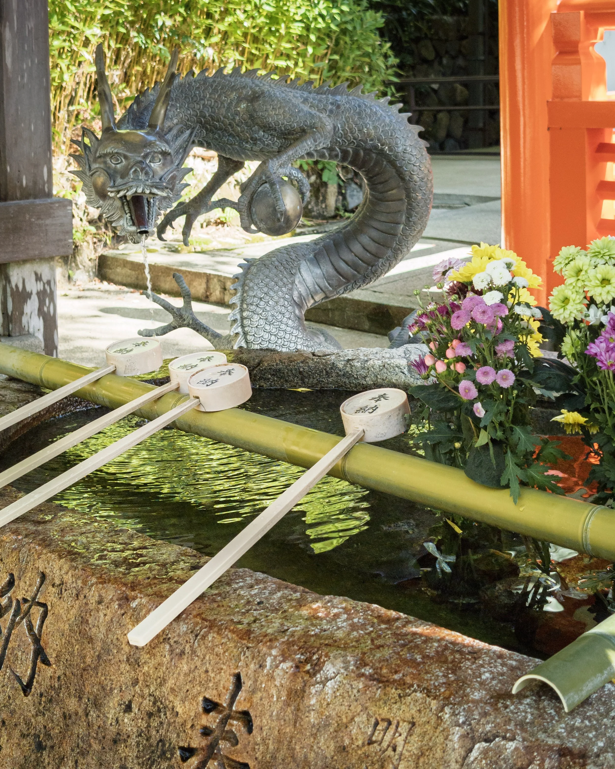 A metal sculpture of a dragon with an elongated body and a fierce expression, sitting beside a small water feature. The sculpture has detailed scales, prominent horns, and an open mouth with water flowing out. There are vibrant flowers and a bamboo water spout in the scene, with greenery in the background.